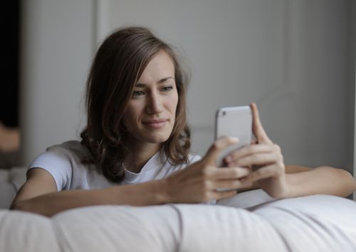 Free Woman comfortably sitting on a couch using her smartphone with a smile, enjoying leisure time indoors. Stock Photo