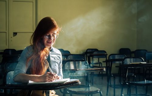 Free A young woman with glasses writes in a sunlit classroom, creating a serene study atmosphere. Stock Photo