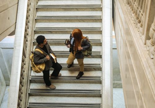 Free Two students with backpacks and books talking on a marble staircase indoors. Stock Photo