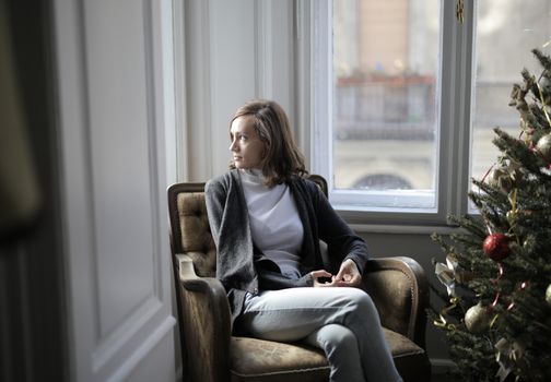 Woman sitting in an armchair by a Christmas tree, gazing out the window with a contemplative look.