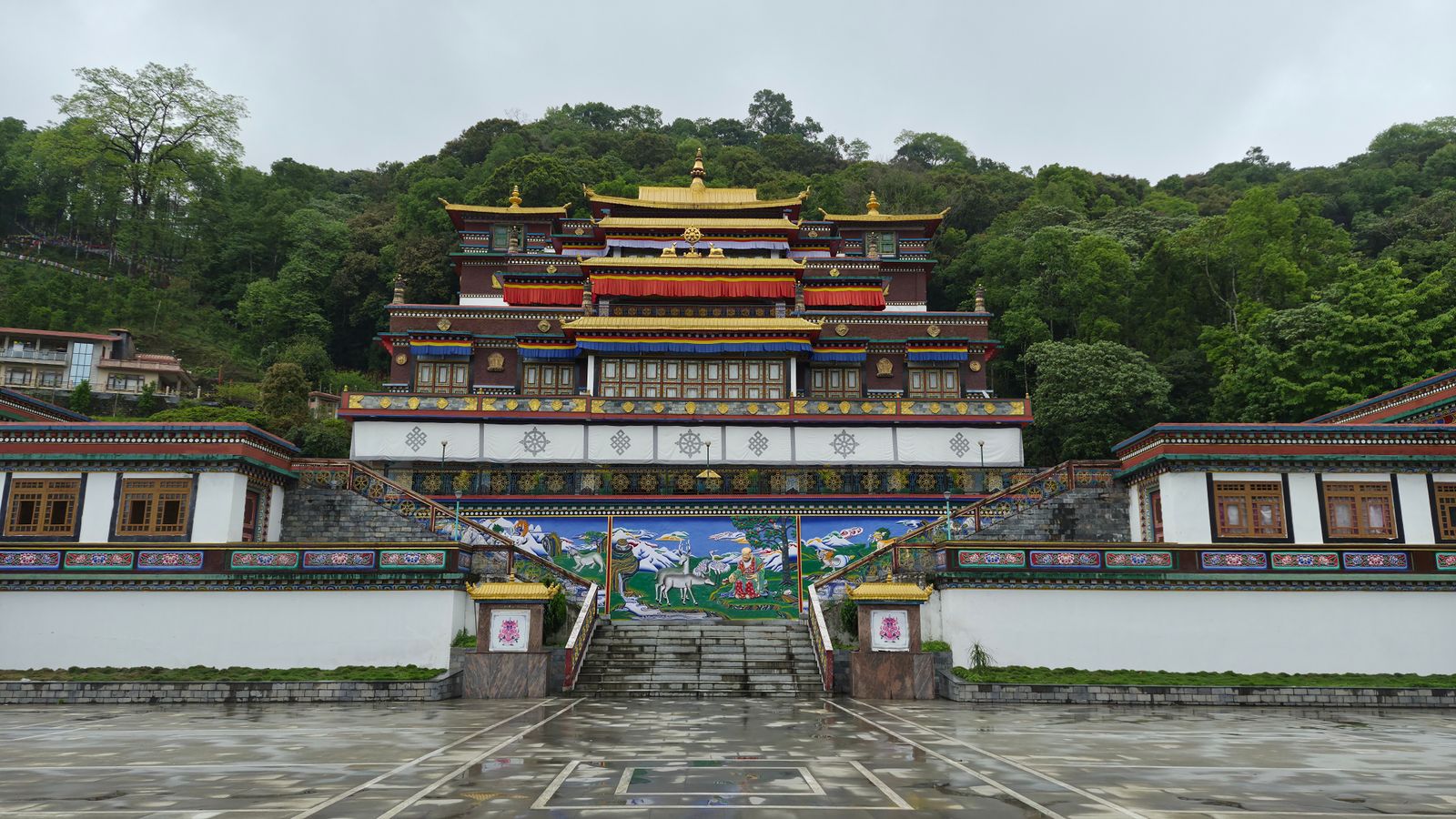 The main shrine of Rumtek Monastery above its rain washed courtyard, gilded roofs and a painted stair wall against forest