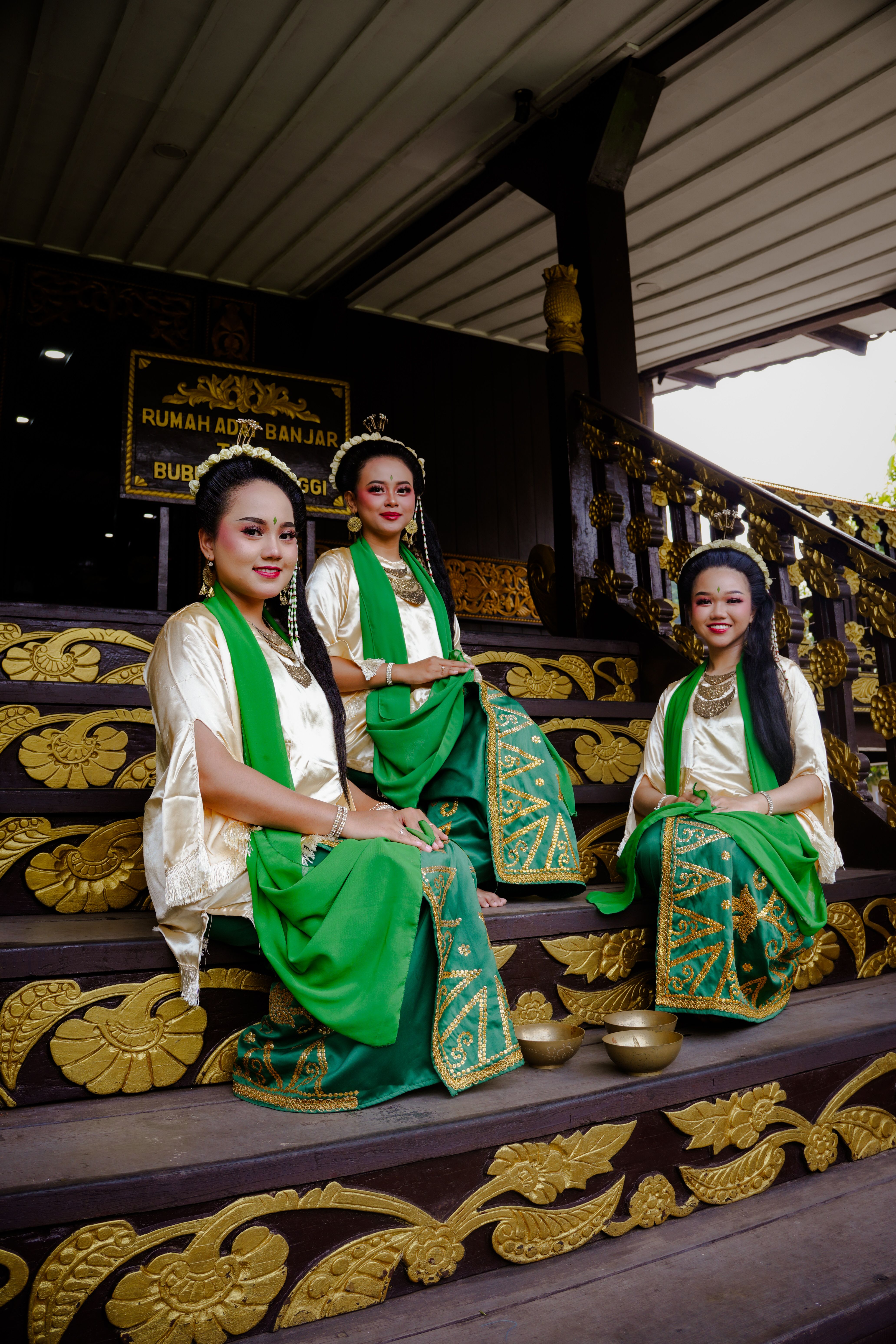 Three women in traditional Banjarese attire seated on decorative steps with cultural backdrop.