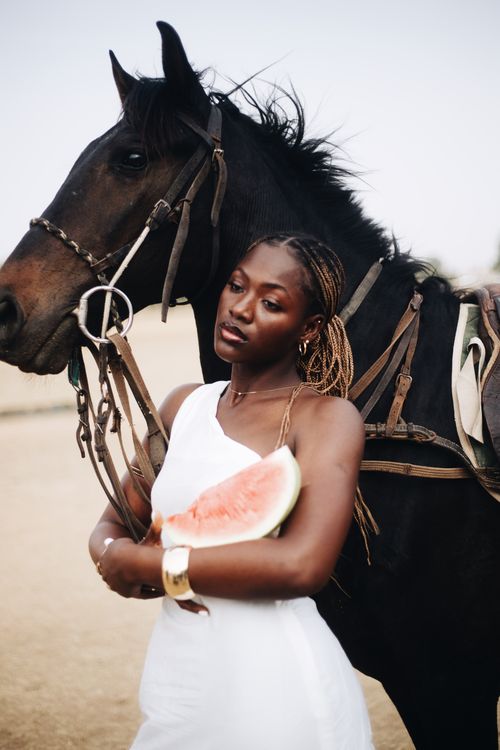 Free A woman in a white dress holds a watermelon beside a black horse, outdoors. Stock Photo