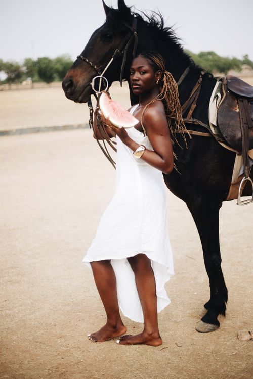 Free A woman in a white dress stands with a horse holding a slice of watermelon outdoors. Stock Photo