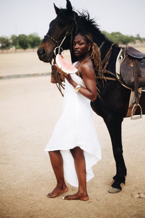 Free Woman in white dress holding watermelon beside a horse outdoors. Stock Photo