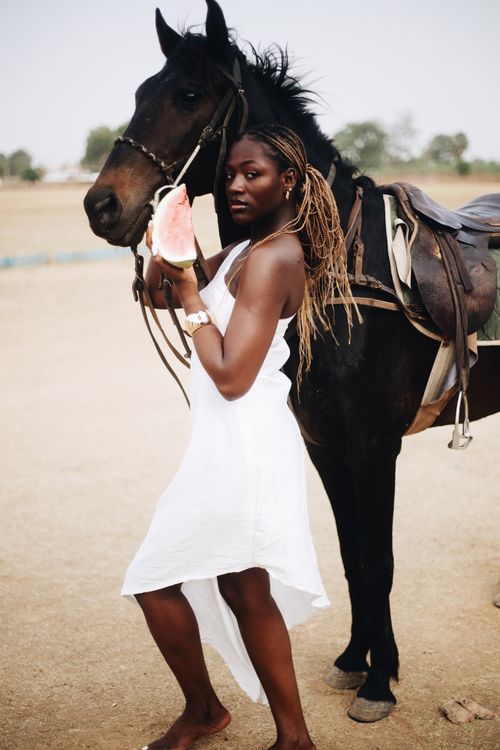 Free A woman in a white dress holds a watermelon slice standing beside a black horse outdoors. Stock Photo