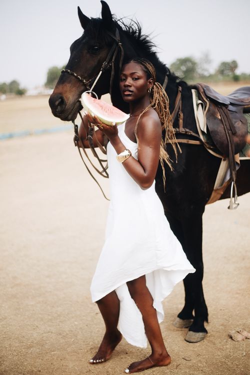 Free A woman in a white dress standing with a horse, holding a watermelon slice outdoors. Stock Photo