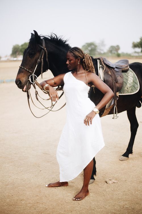 Free A woman in a white dress stands stylishly beside a horse on a sunny day. Stock Photo