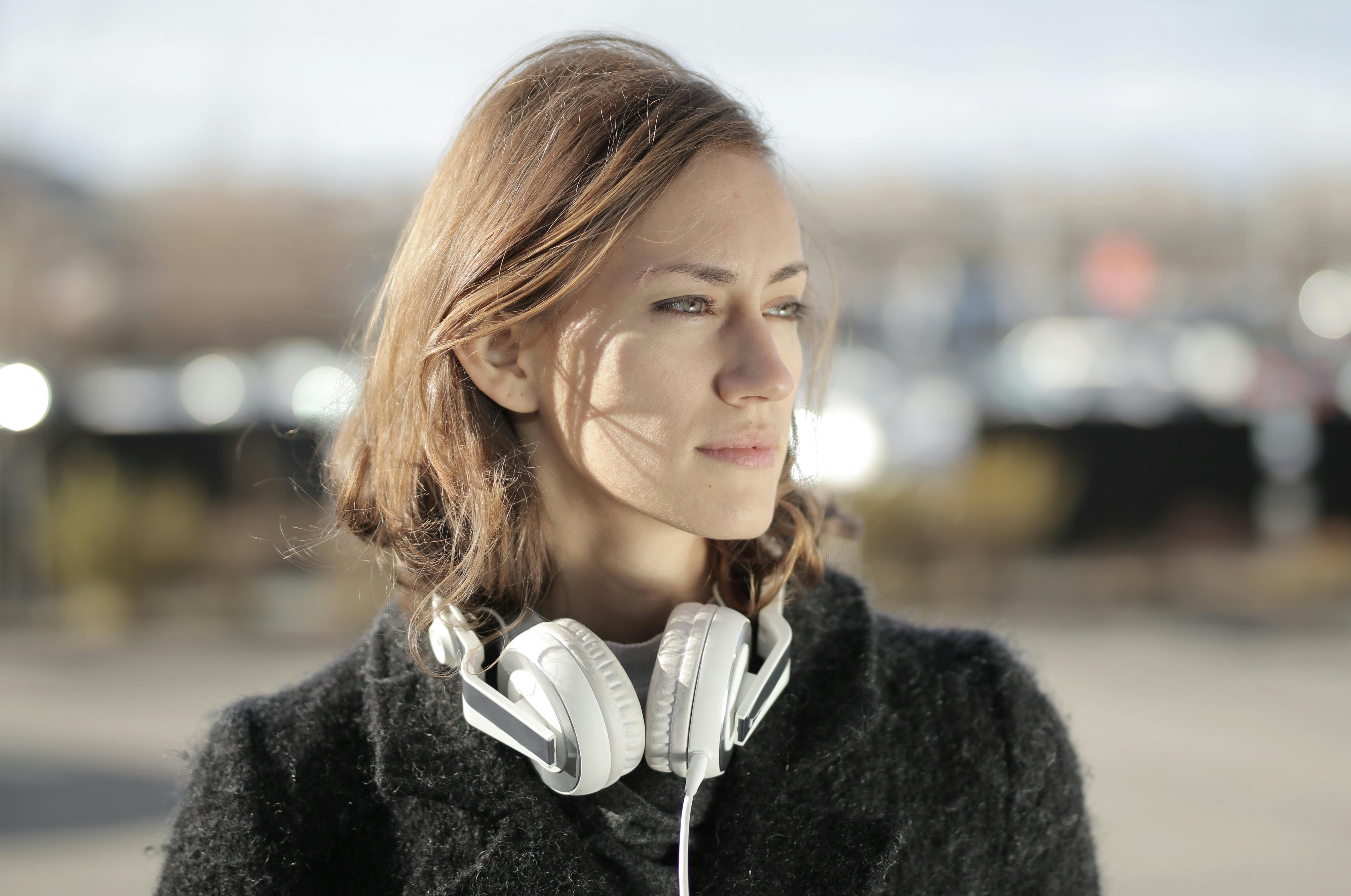 A woman in a fur coat with headphones, gazing thoughtfully outdoors in soft daylight.