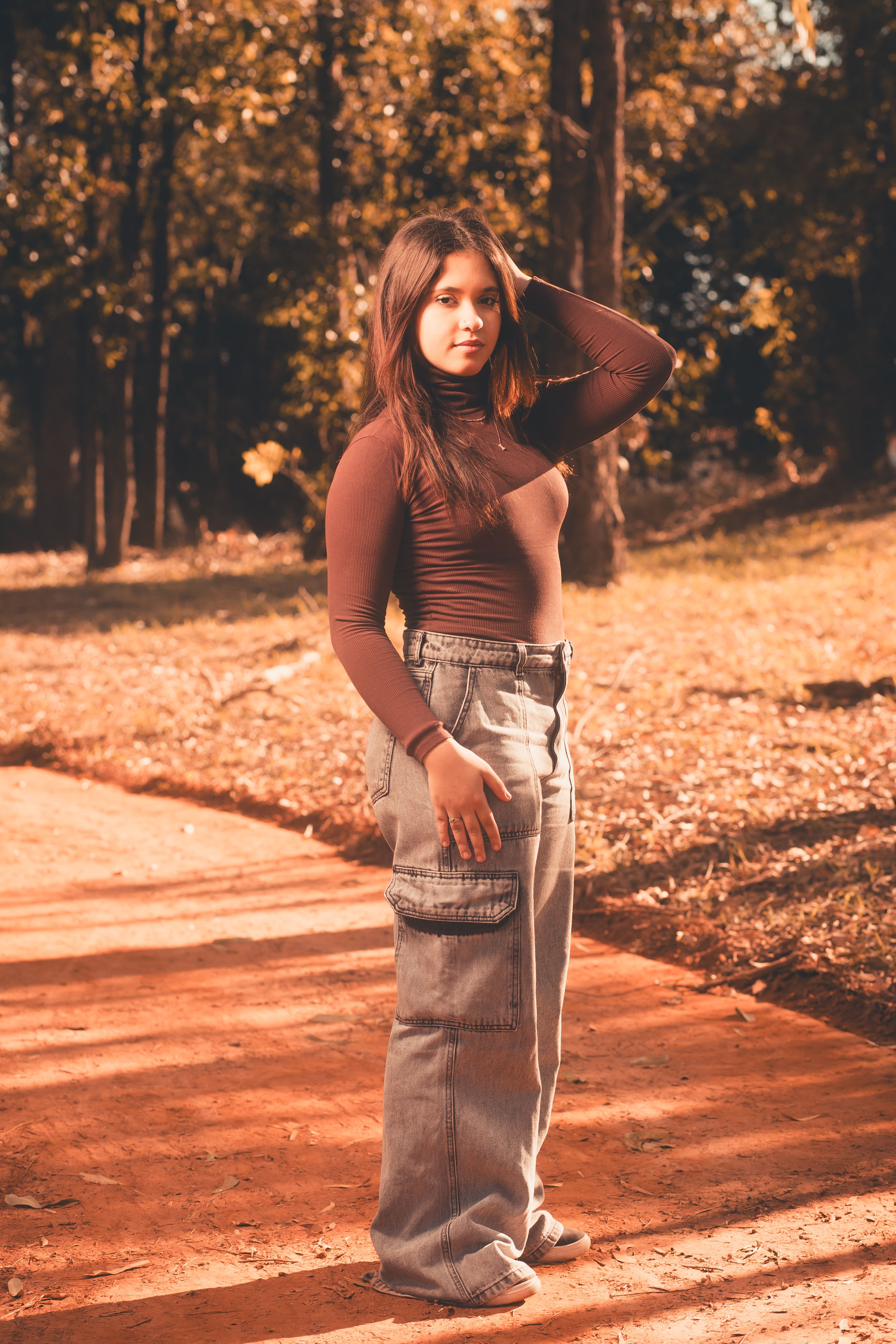 Stylish woman in autumn outfit poses on a sunlit park pathway surrounded by vibrant fall foliage.