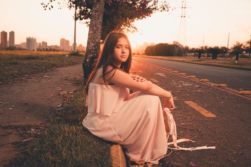 Free A young woman in a flowing dress sits by a city road at sunset, capturing a serene moment. Stock Photo