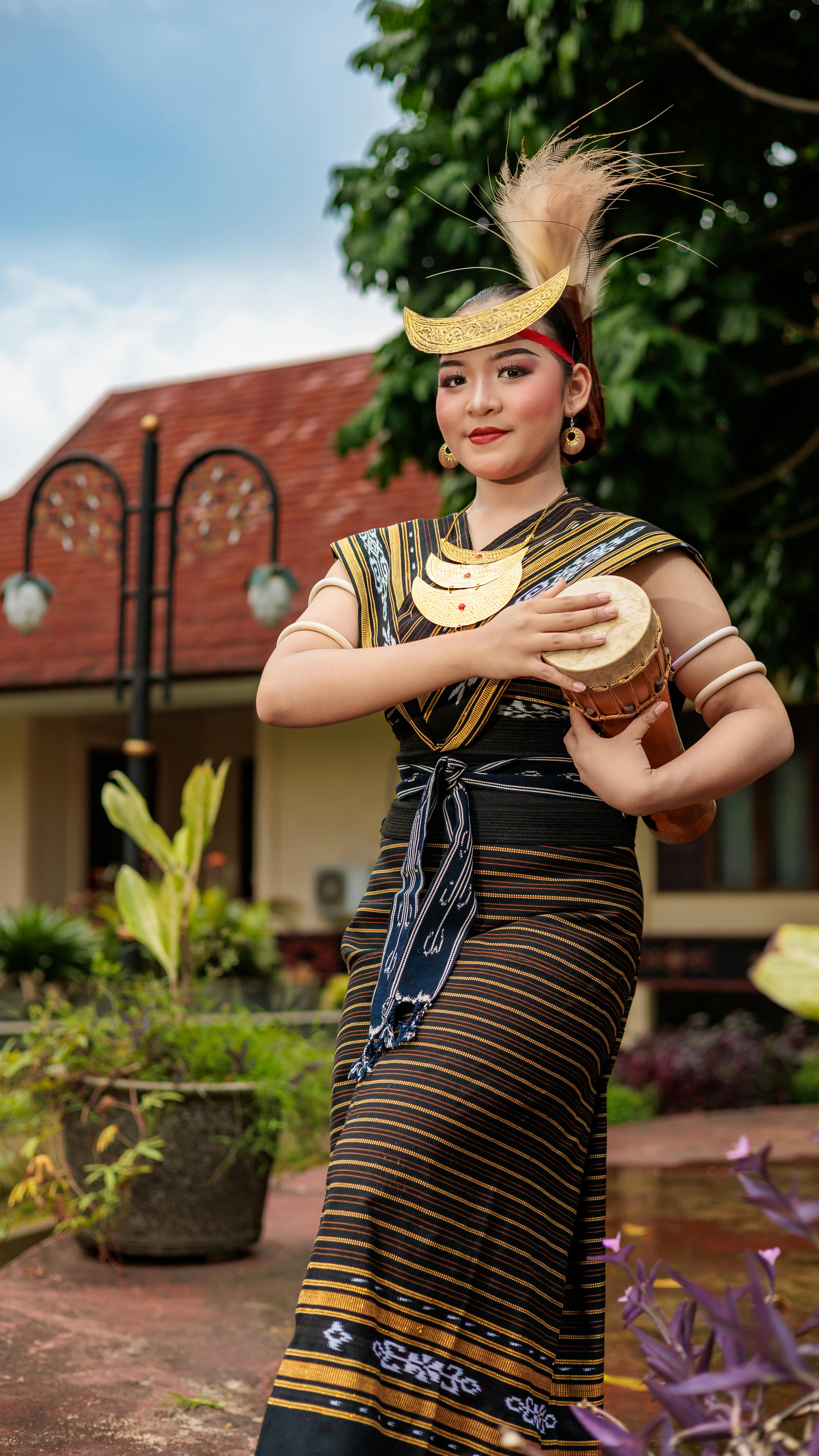 Young woman in ethnic attire plays a hand drum outdoors, showcasing cultural heritage.