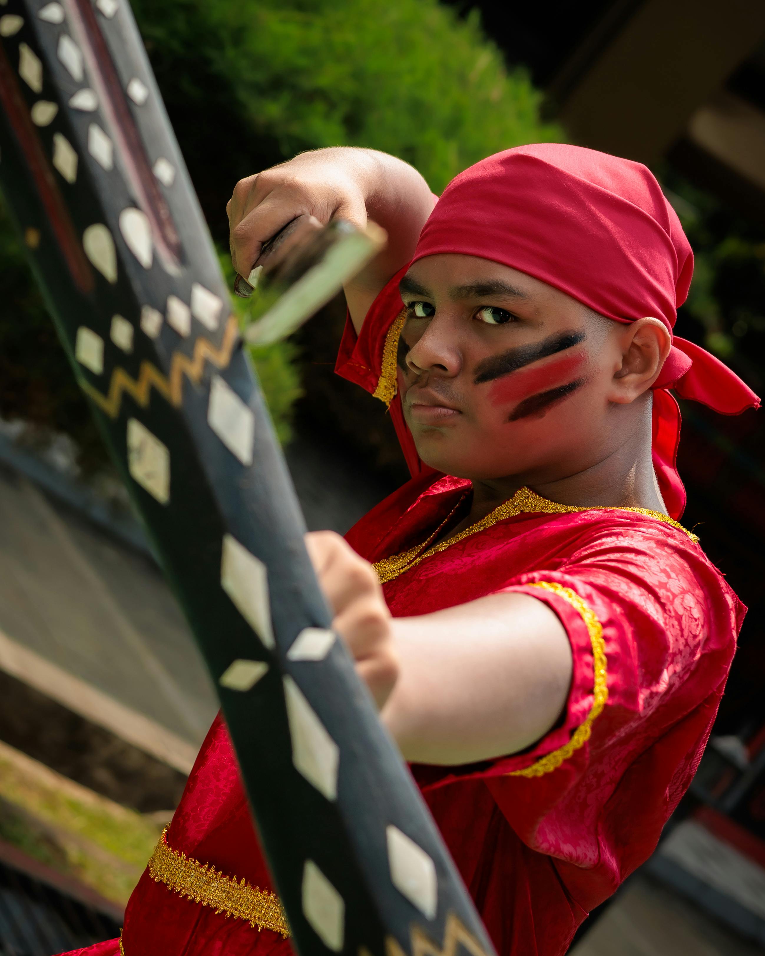 A young boy in traditional red attire strikes a pose outdoors, holding a decorated weapon.