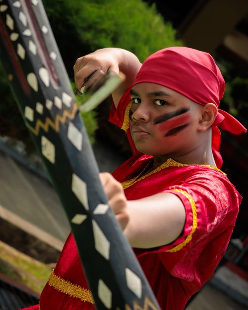 Free A young boy in traditional red attire strikes a pose outdoors, holding a decorated weapon. Stock Photo