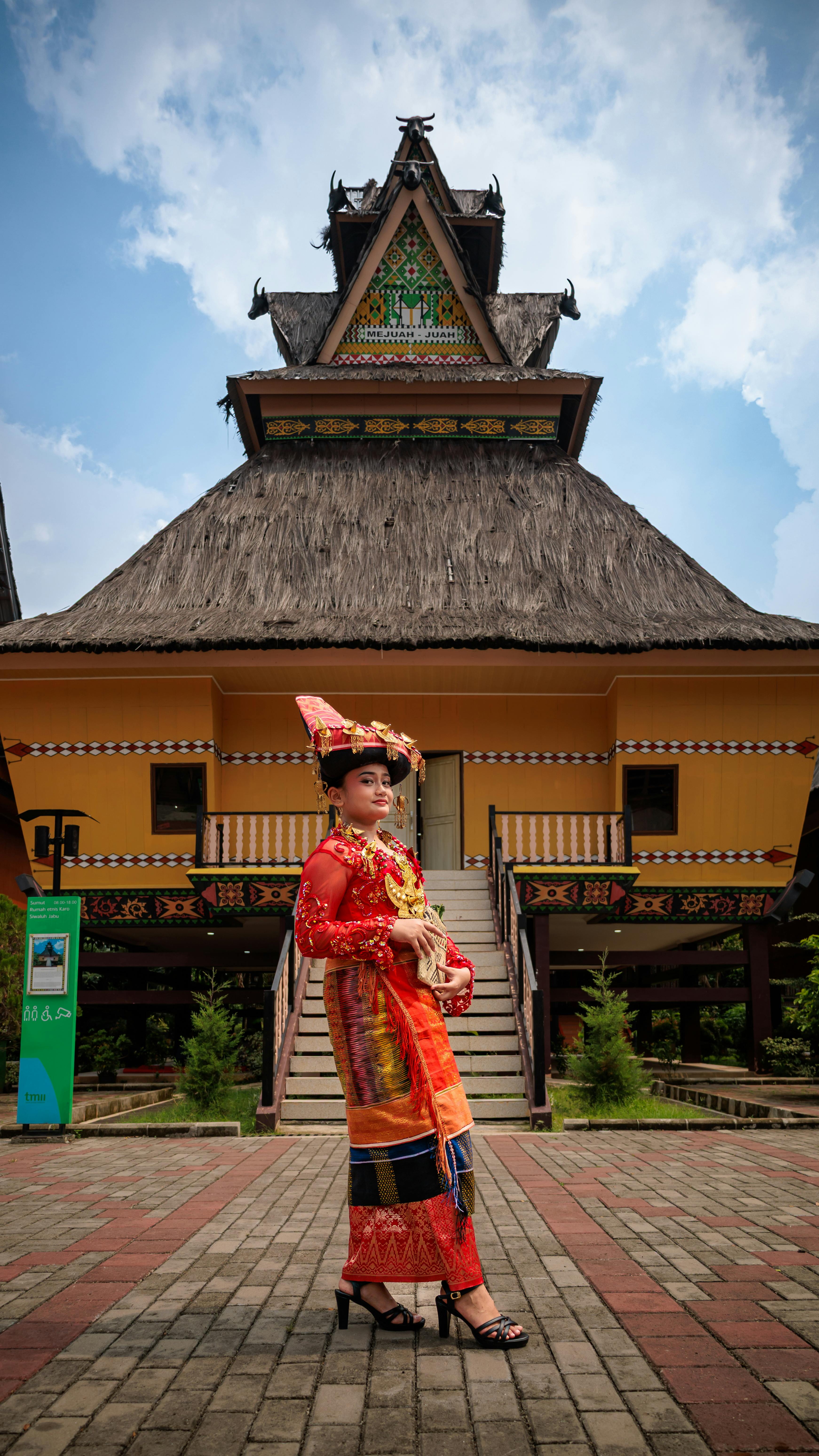 Woman in traditional Batak attire poses in front of iconic Batak house in Jakarta, Indonesia.