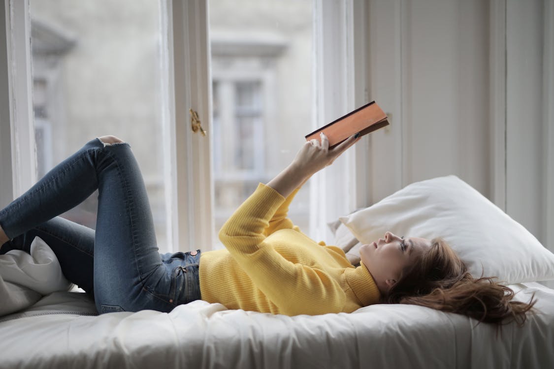 Free Woman in Yellow Sweater Lying on Bed While Reading Book Stock Photo