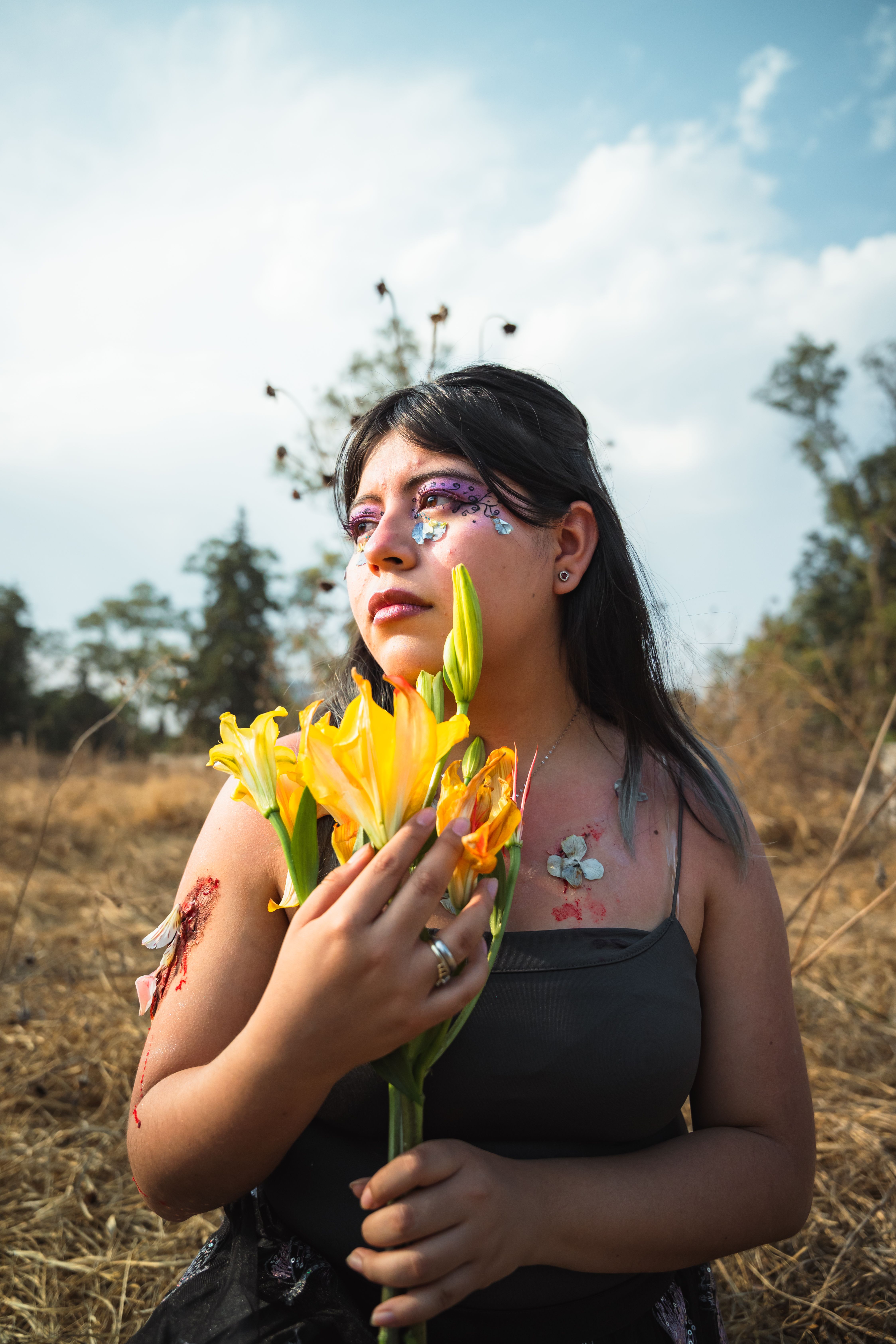 Fotomural Una mujer con maquillaje artístico sostiene flores amarillas en un campo soleado y seco.