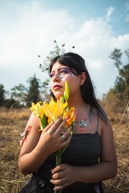Fotomural Una mujer con maquillaje artístico sostiene flores amarillas en un campo soleado y seco.