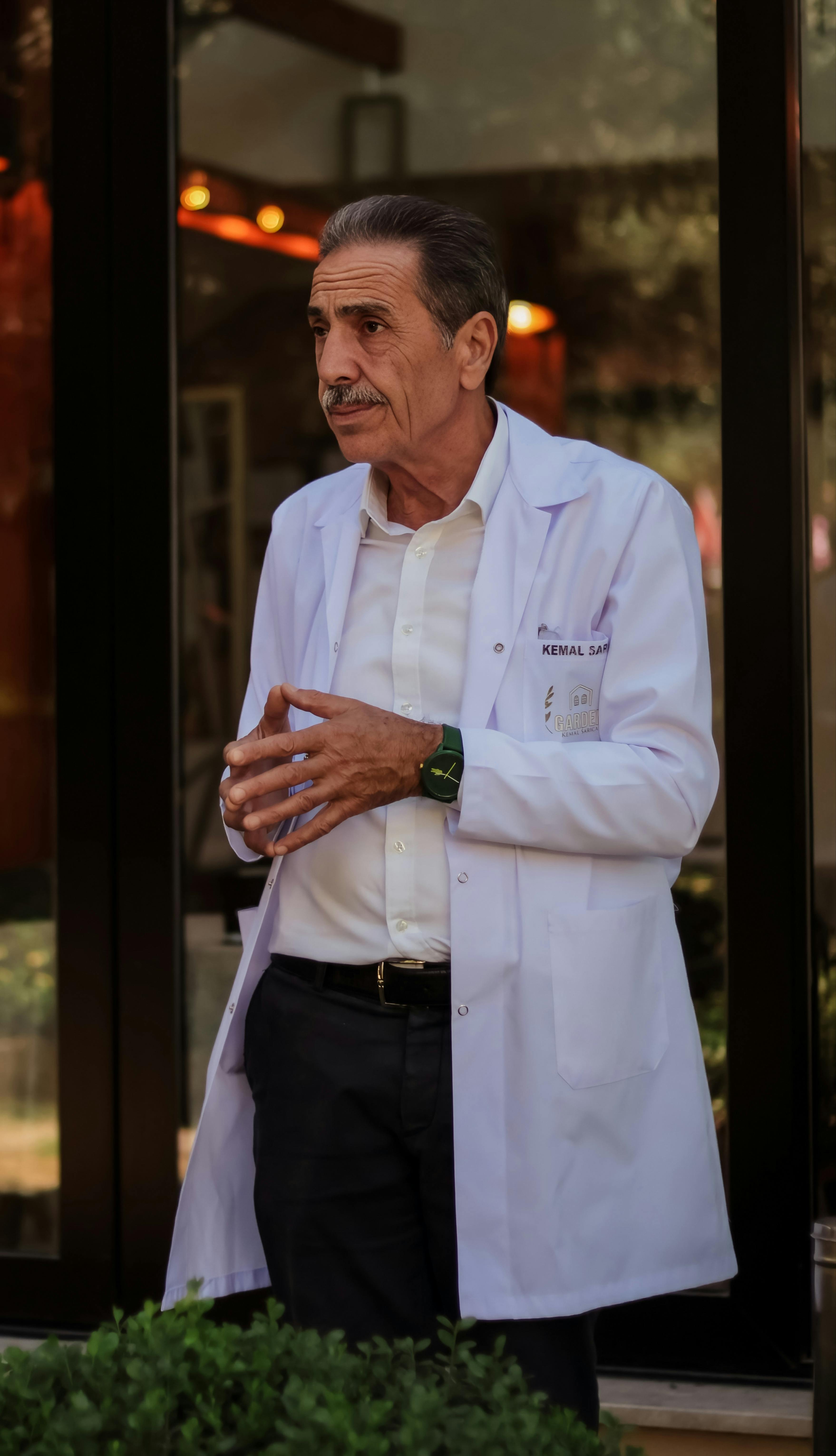A senior doctor standing in front of a building, wearing a white coat and engaging in conversation.