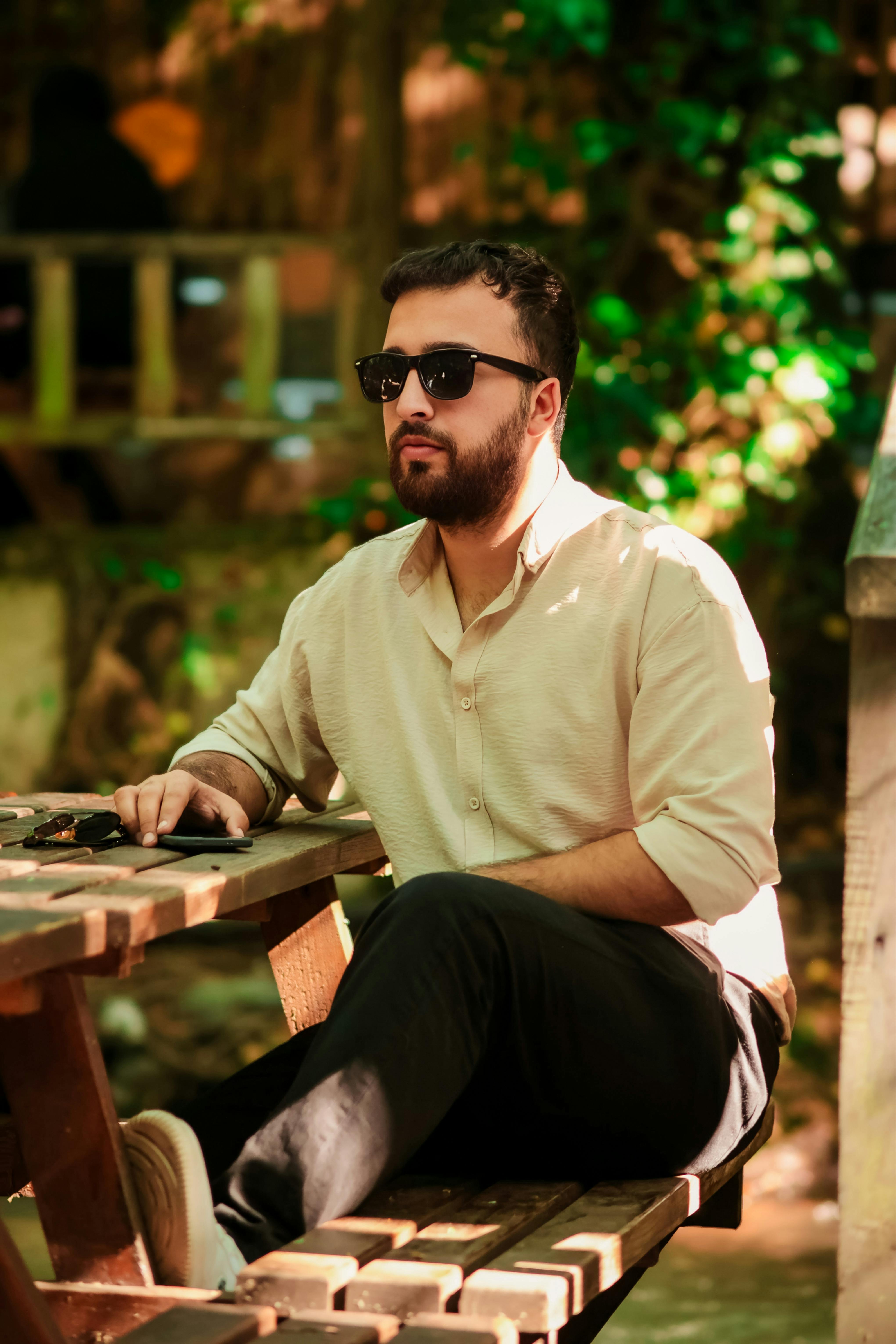 Stylish man wearing sunglasses sitting at a wooden table outdoors in a sunny cafe.