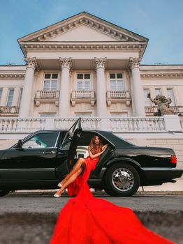 A glamorous woman in a flowing red dress poses by a classic car in front of a grand building.
