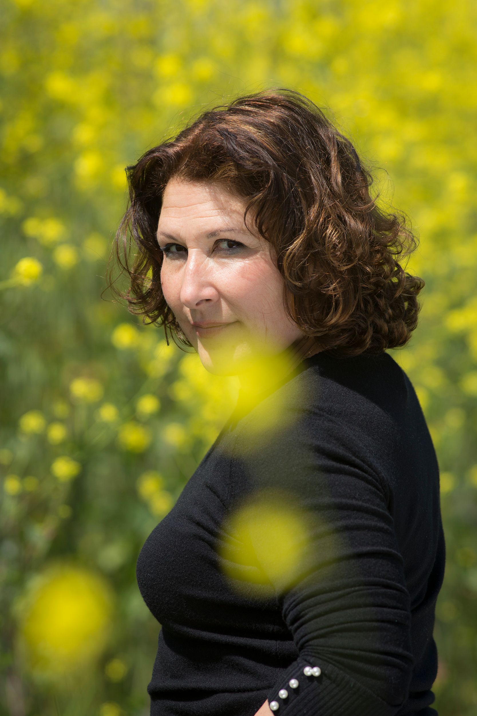 A woman in a black outfit stands amidst a vibrant yellow wildflower field, looking back.