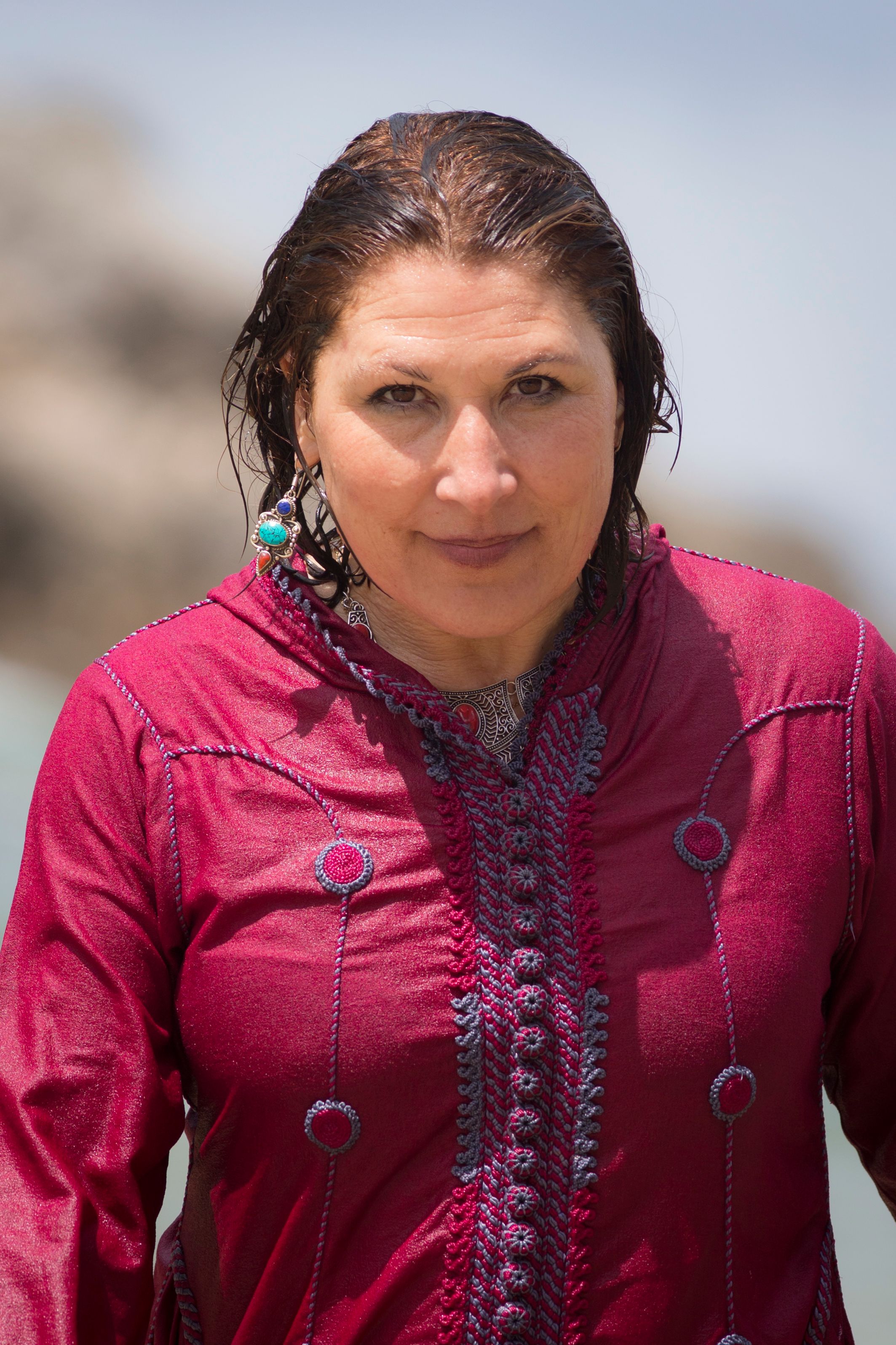 A woman in vibrant clothing poses confidently outdoors with a blurred scenic background.