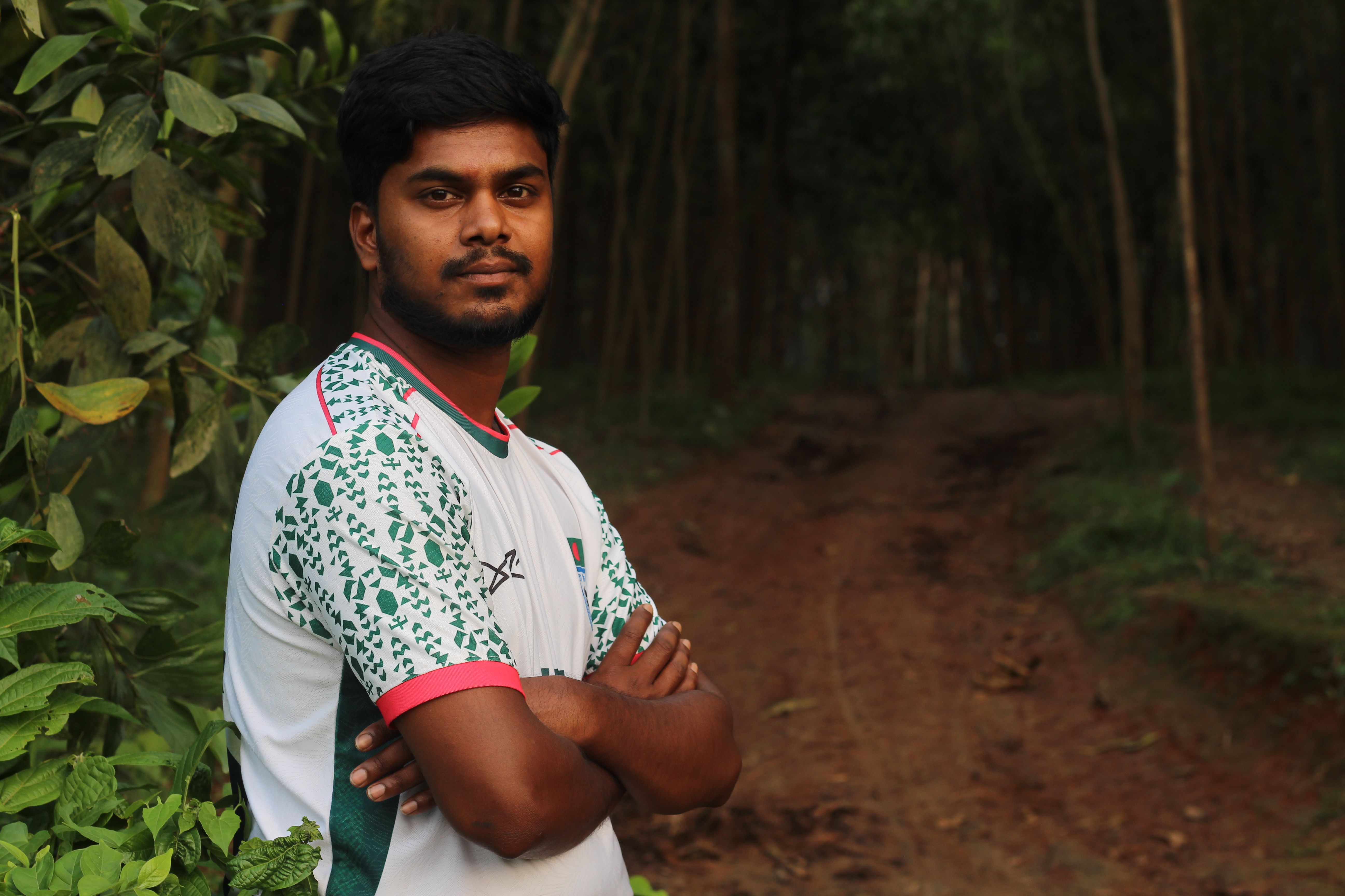 Young man standing confidently on a forested path, surrounded by greenery.