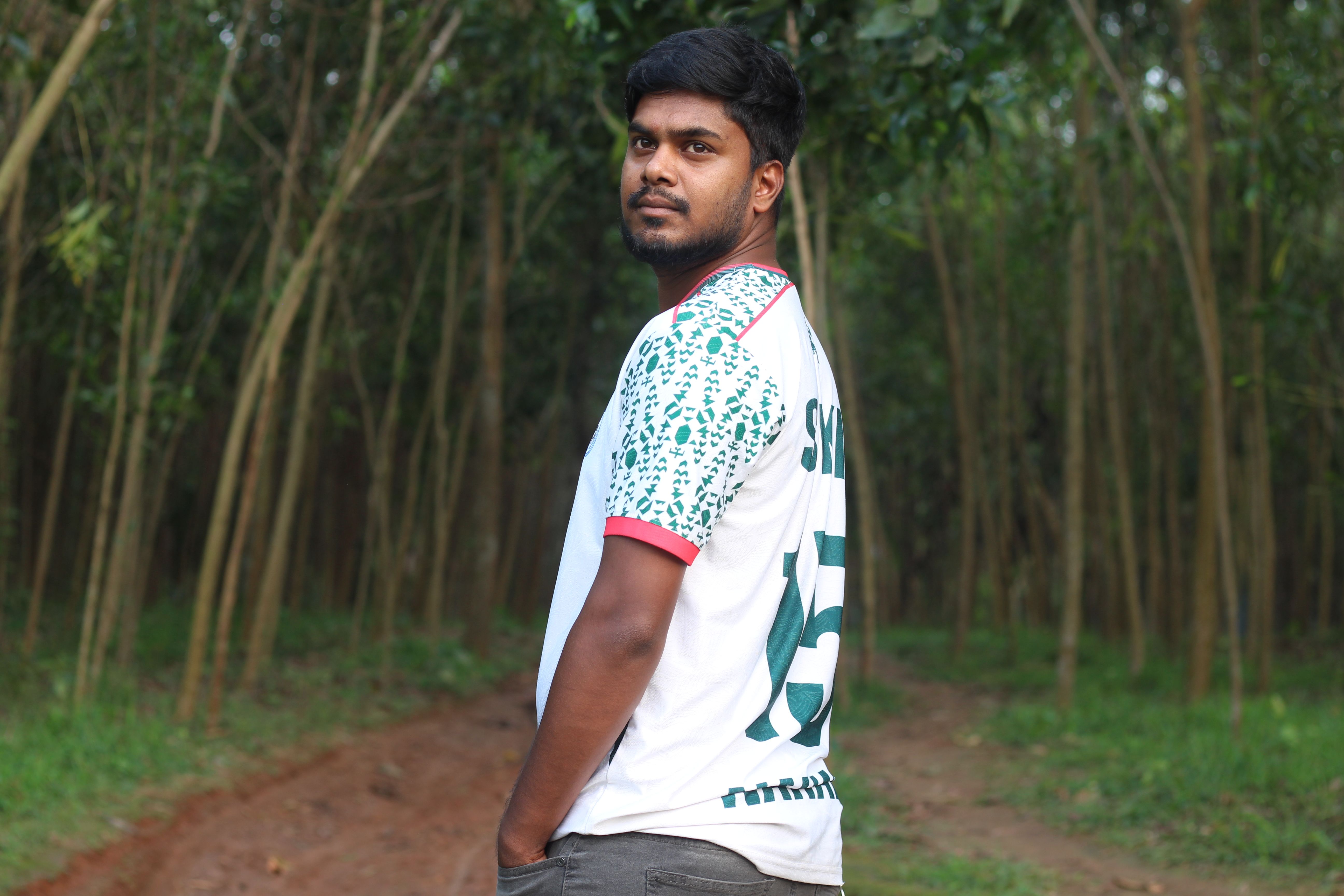 A young man stands confidently on a forest pathway, surrounded by tall trees.