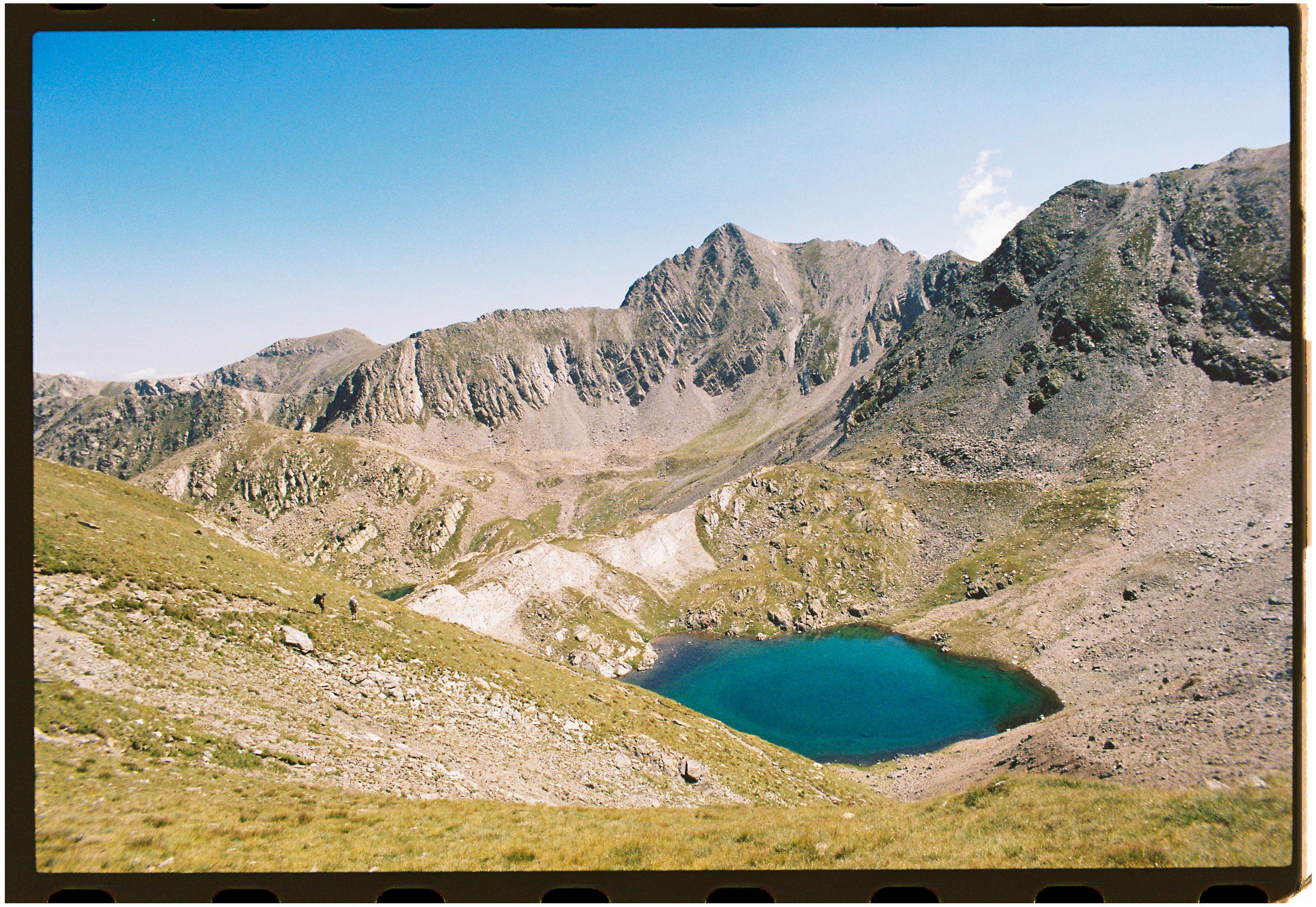 A pristine mountain lake surrounded by rocky and grassy terrain under a clear blue sky.