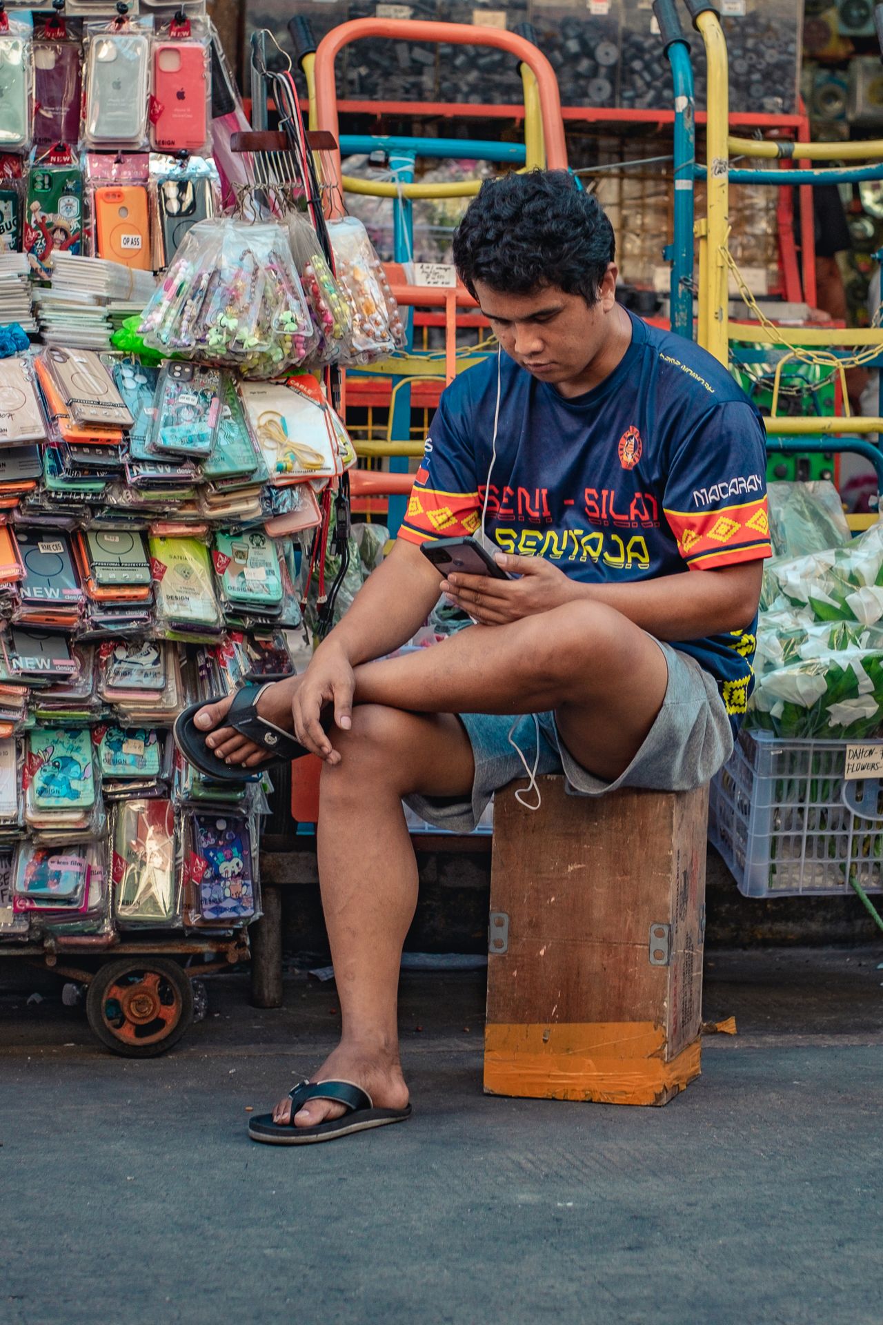 A street vendor in Manila sits among colorful merchandise, focused on his smartphone in a bustling outdoor market.