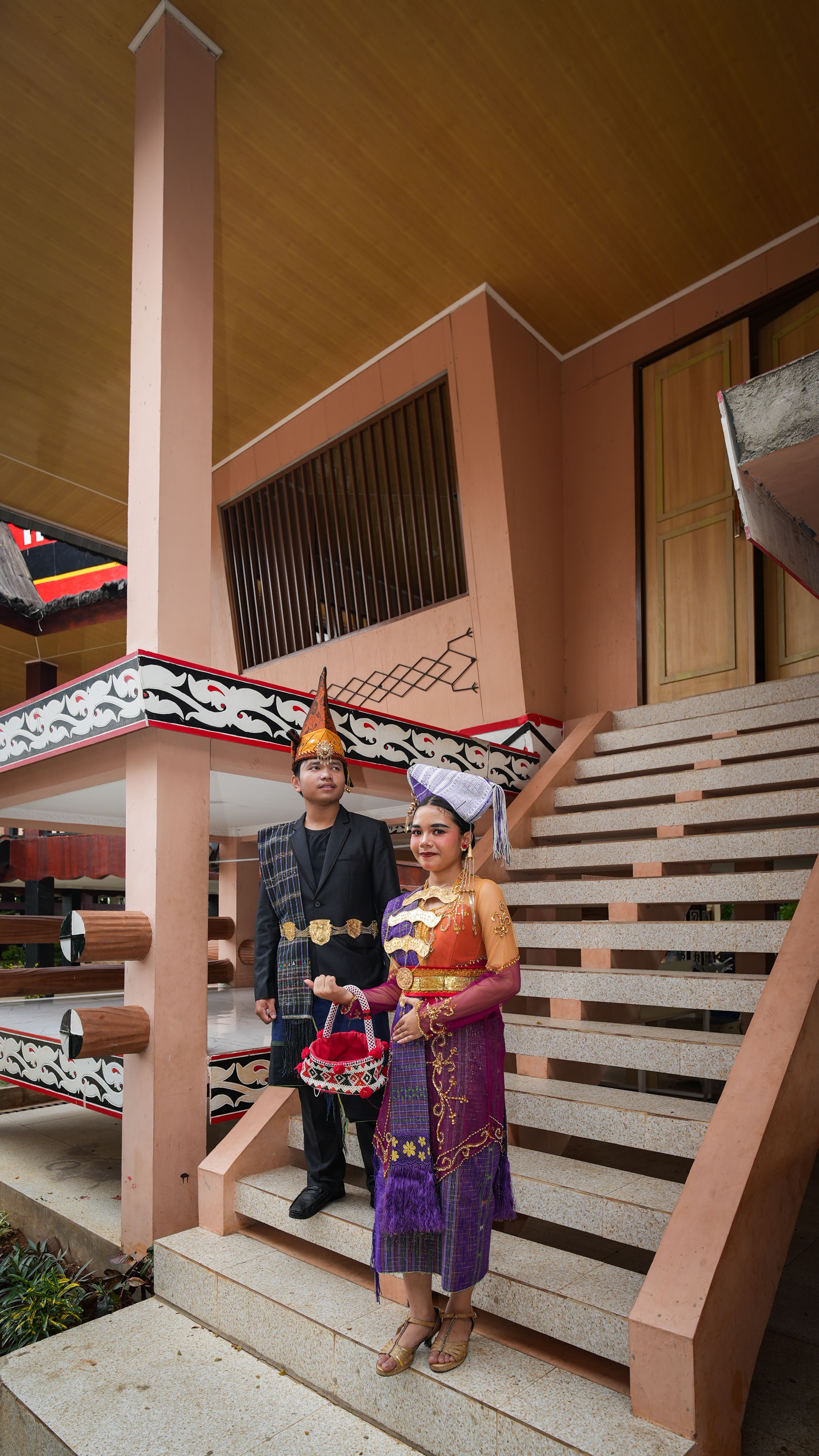 Couple wearing traditional Indonesian clothing on stairs at cultural building.
