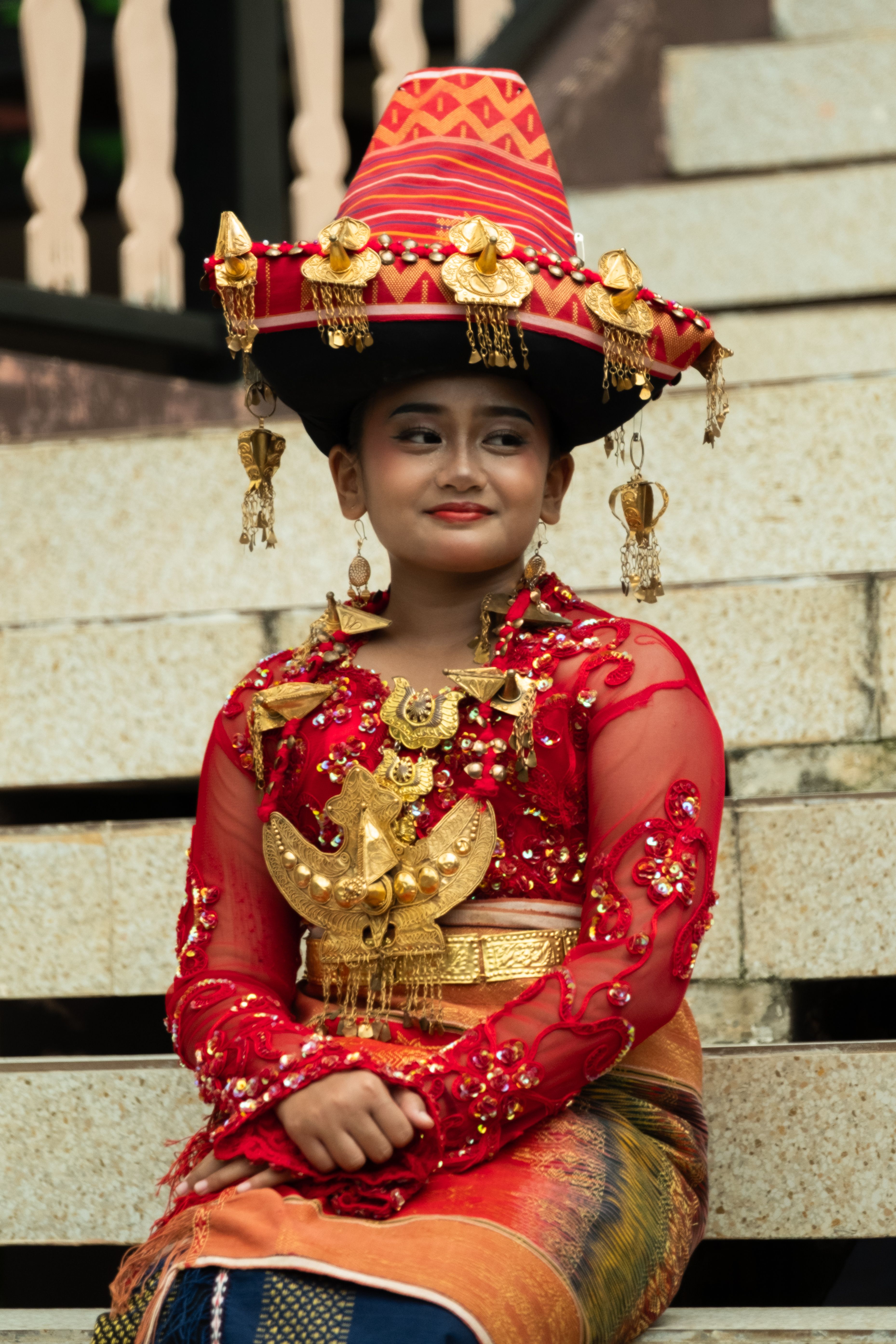 A woman wearing colorful Batak traditional clothing and intricate headwear, sitting on stairs.