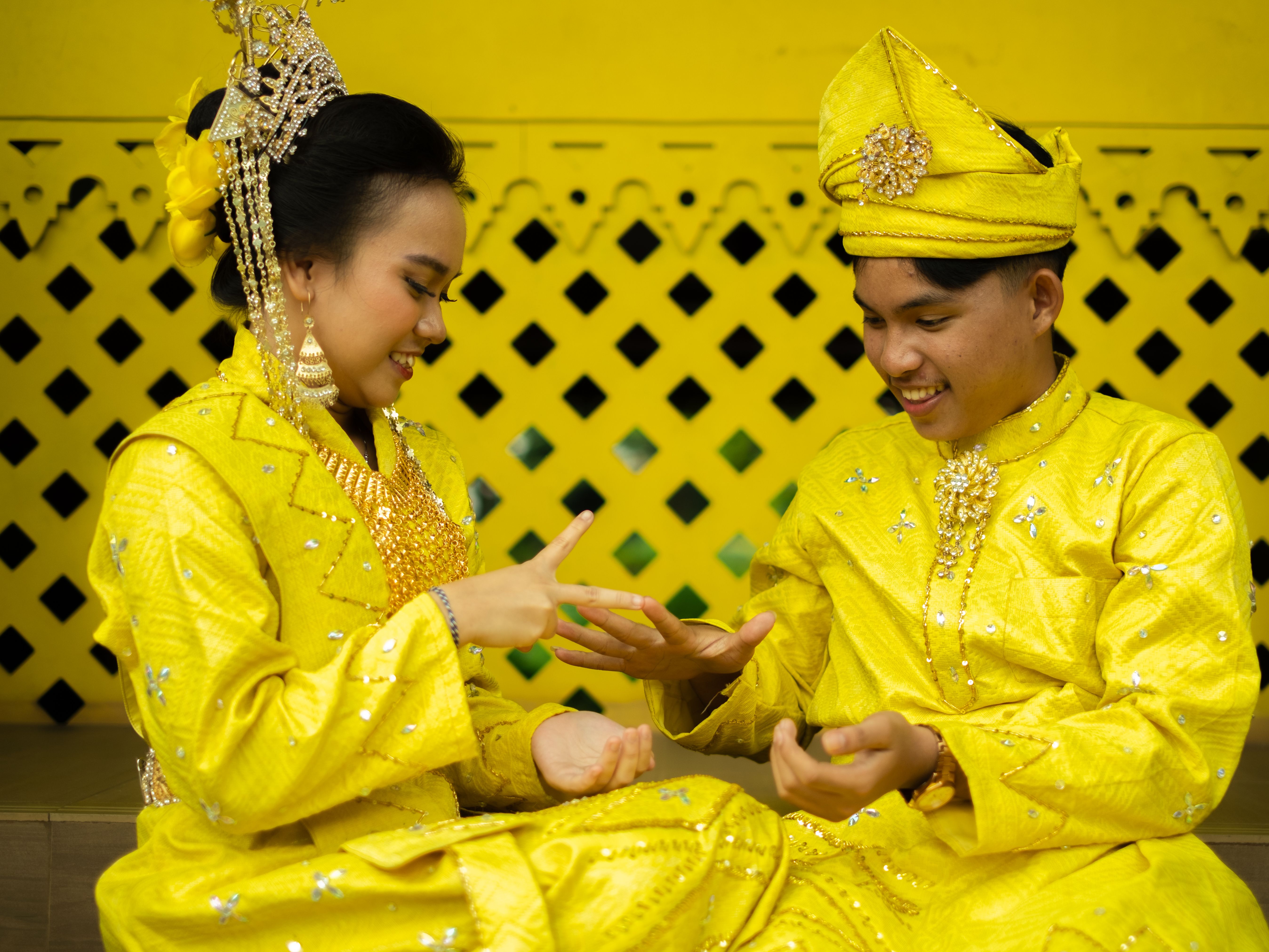 A young couple joyfully playing a game while dressed in vibrant traditional Malay wedding attire.