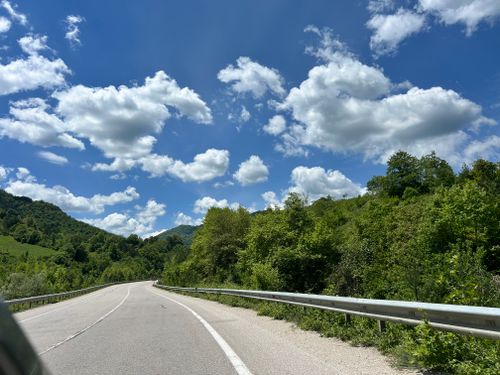 Free A picturesque mountain road with lush greenery and fluffy clouds under a bright blue sky. Stock Photo