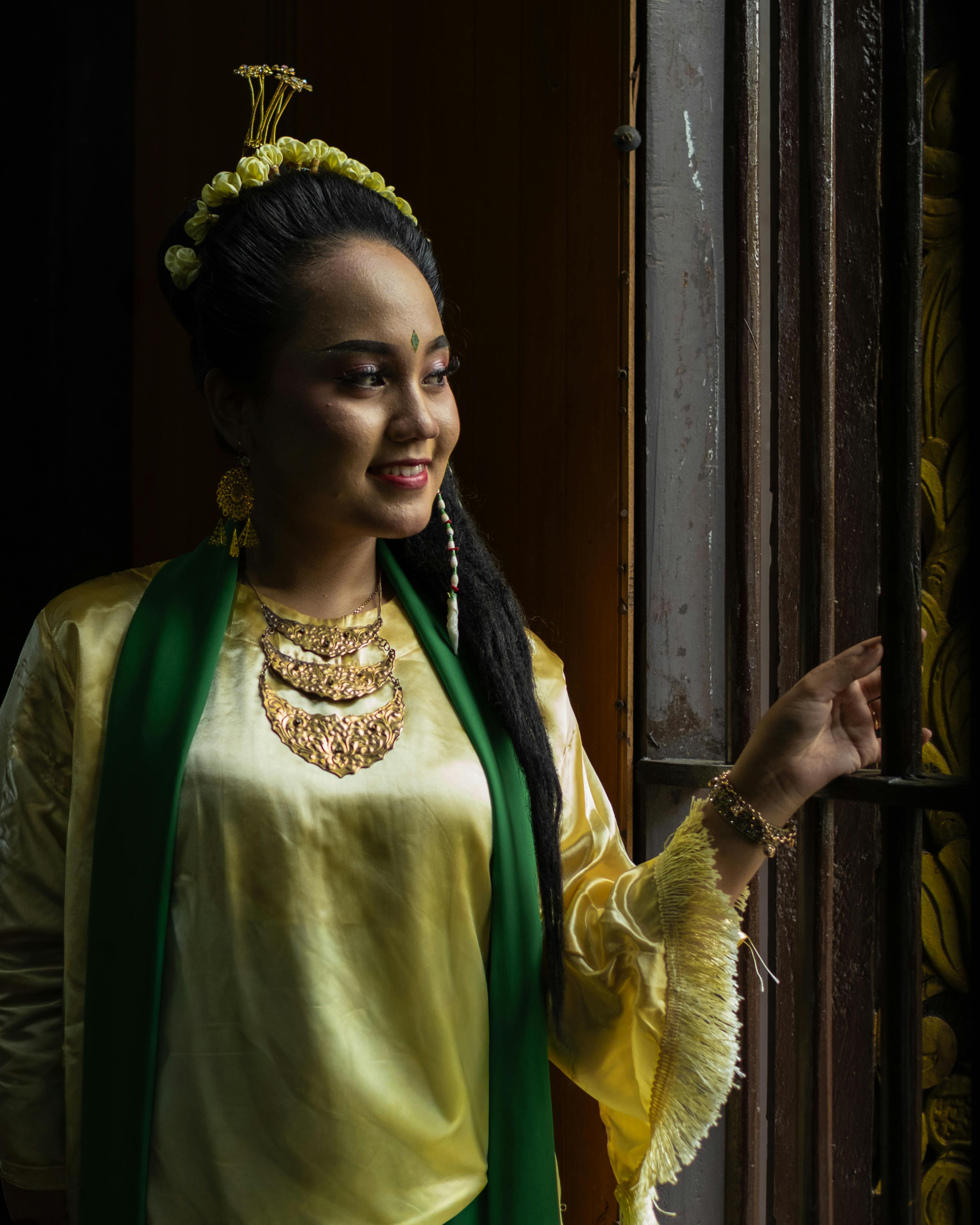 Asian woman in traditional attire and gold jewelry standing by a window with natural light.