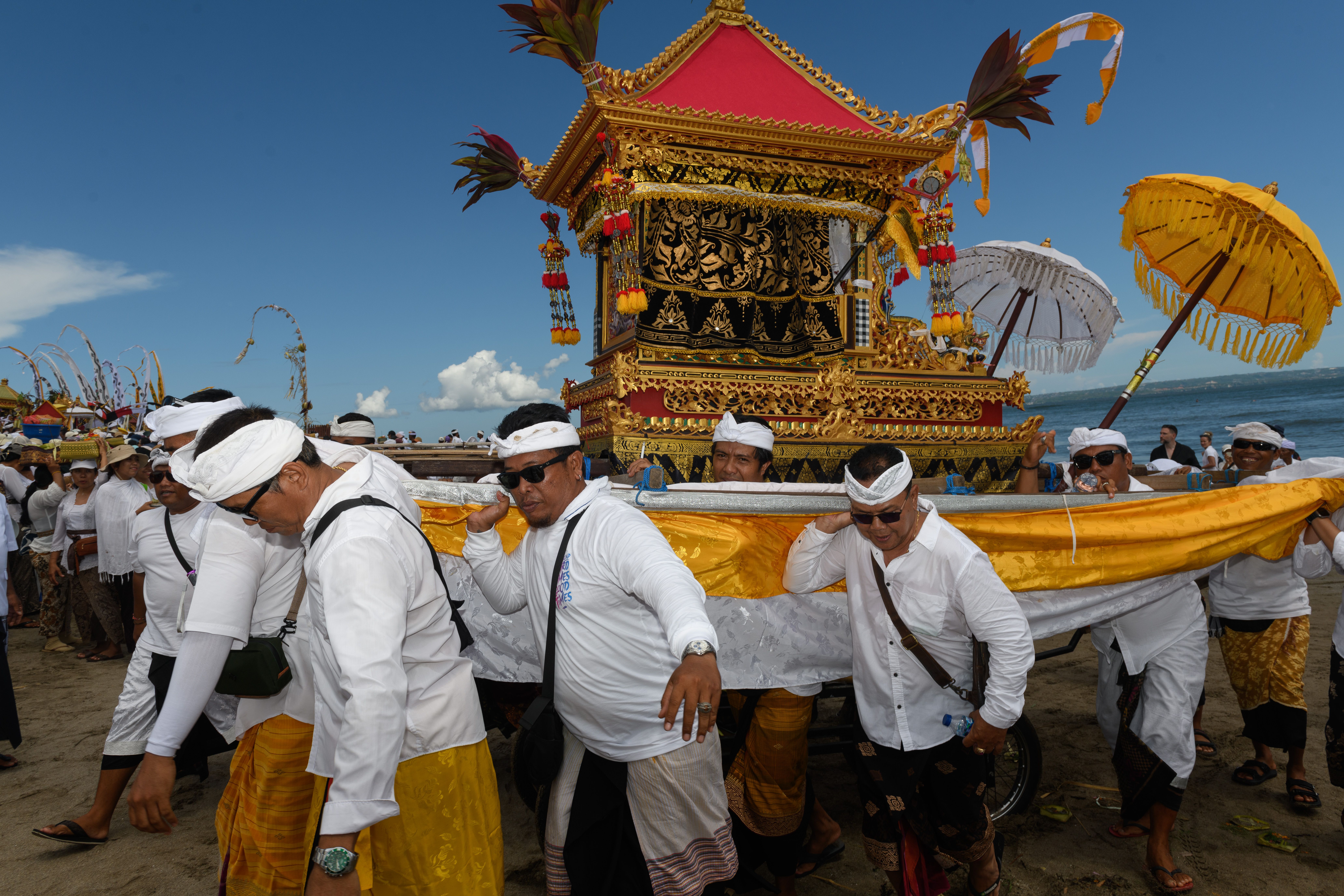 ฟรี ชายชาวบาหลีในชุดพื้นเมืองแบกเกวียนประดับประดาอย่างงดงามในขบวนแห่พิธีริมทะเล คลังภาพถ่าย
