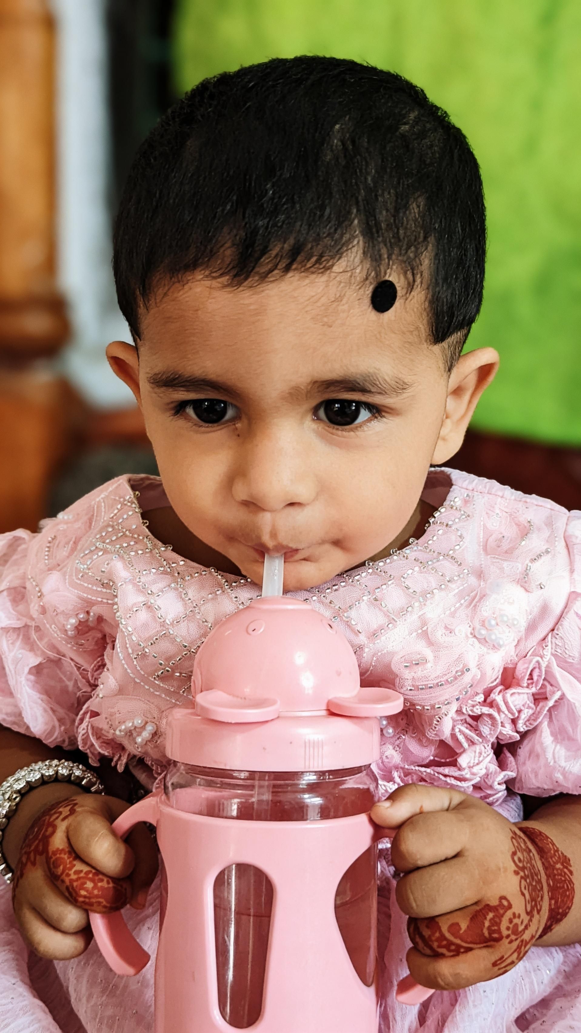 Cute baby in pink dress sipping from a pink cup indoors.