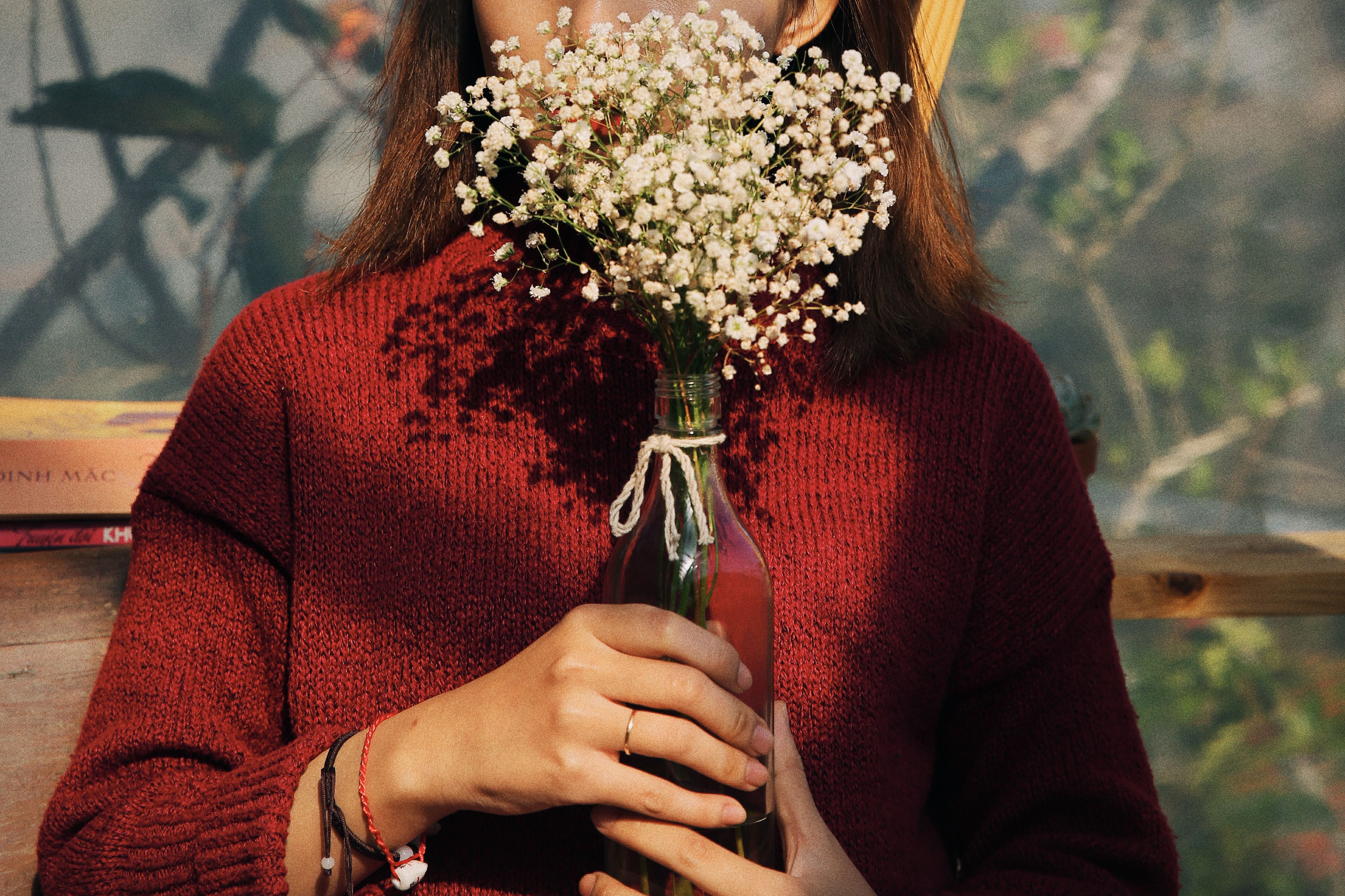 A woman in a red sweater holds a bouquet of baby's breath flowers in a glass bottle.