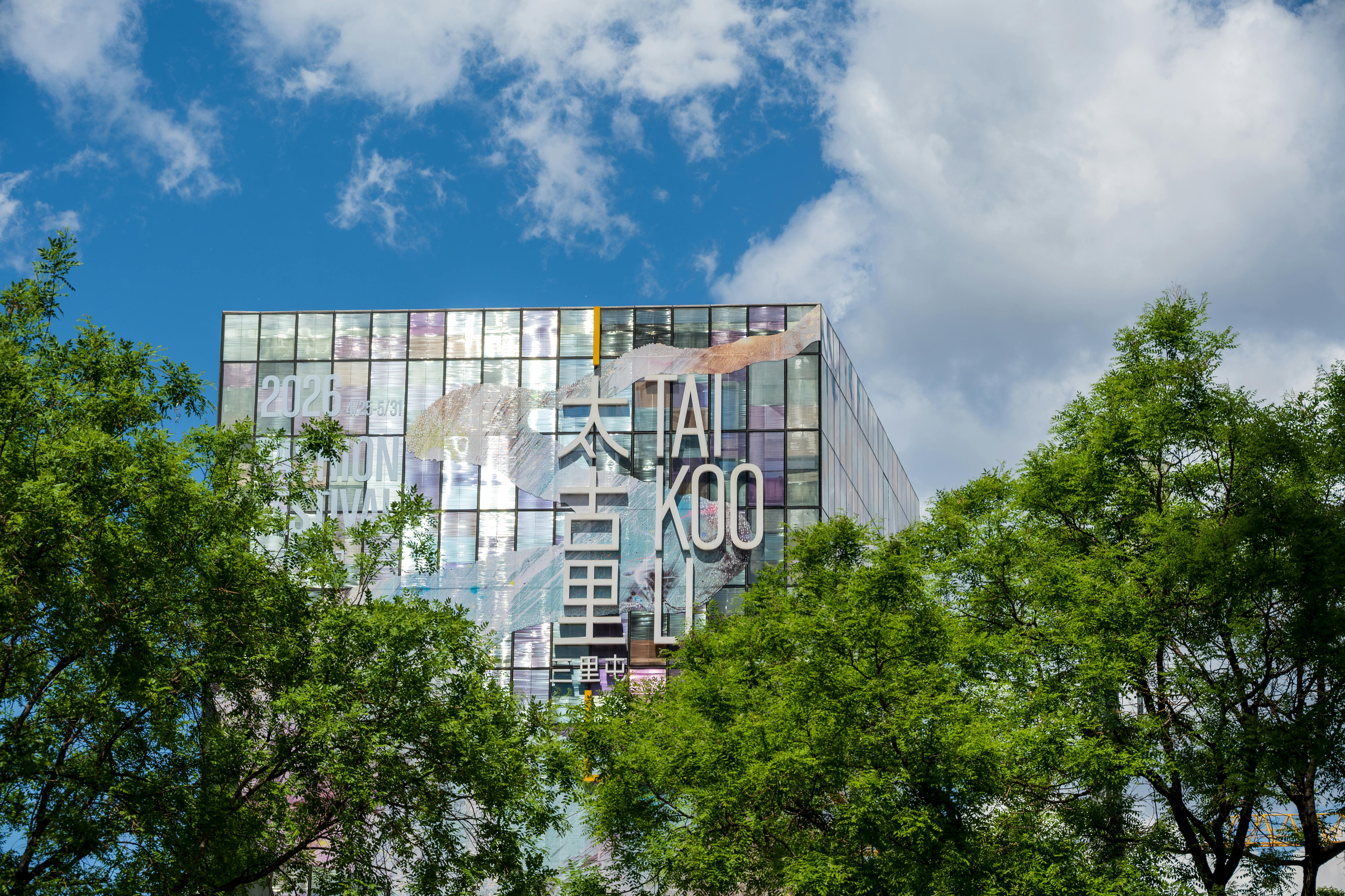 A vibrant cityscape featuring a modern glass building with prominent Tai Koo signage and lush green trees.