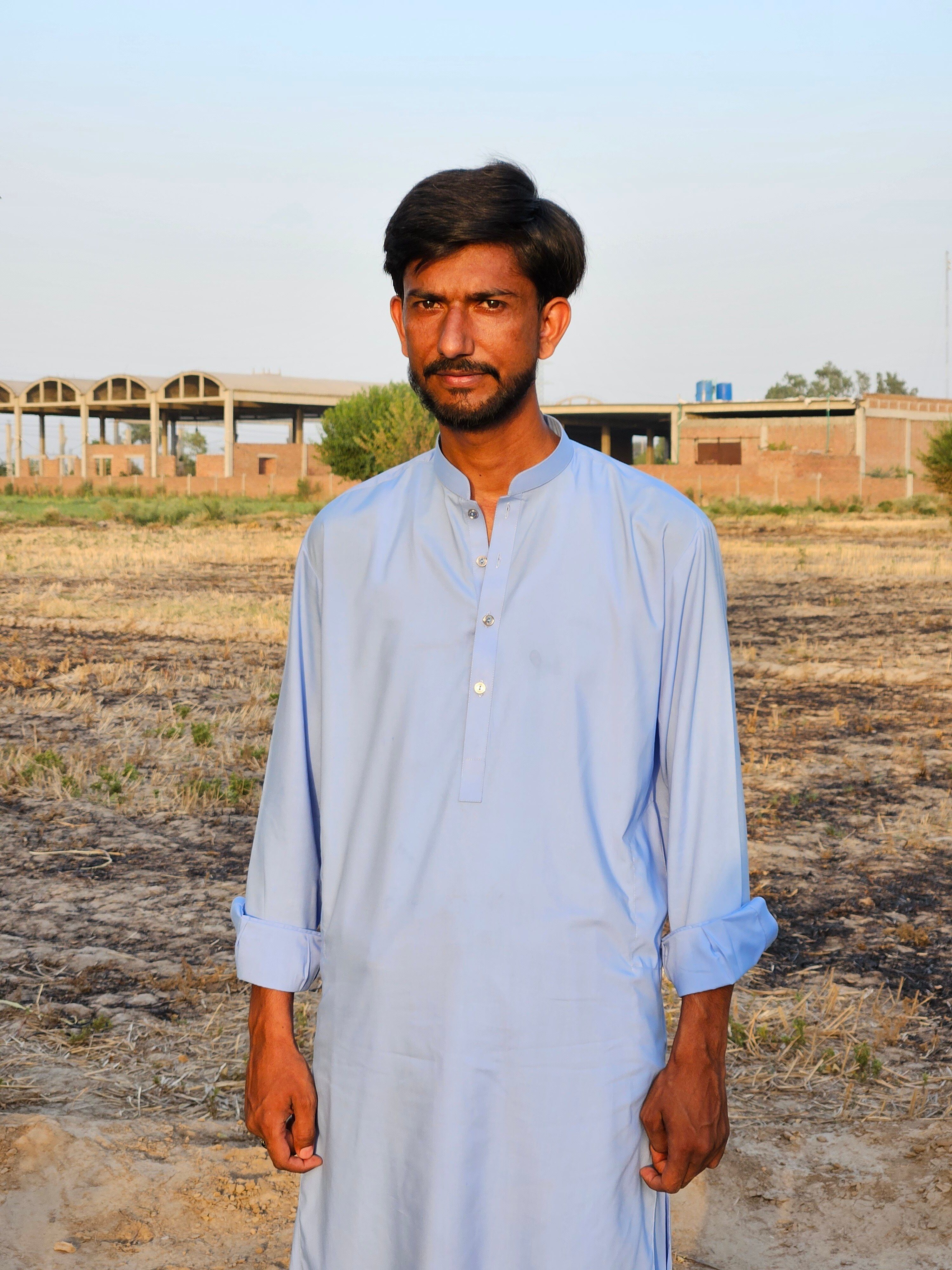 A man in traditional attire stands in a rural outdoor setting during the day.