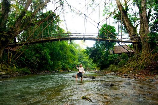 A man crossing a river below a traditional bamboo bridge in lush Banten, Indonesia.