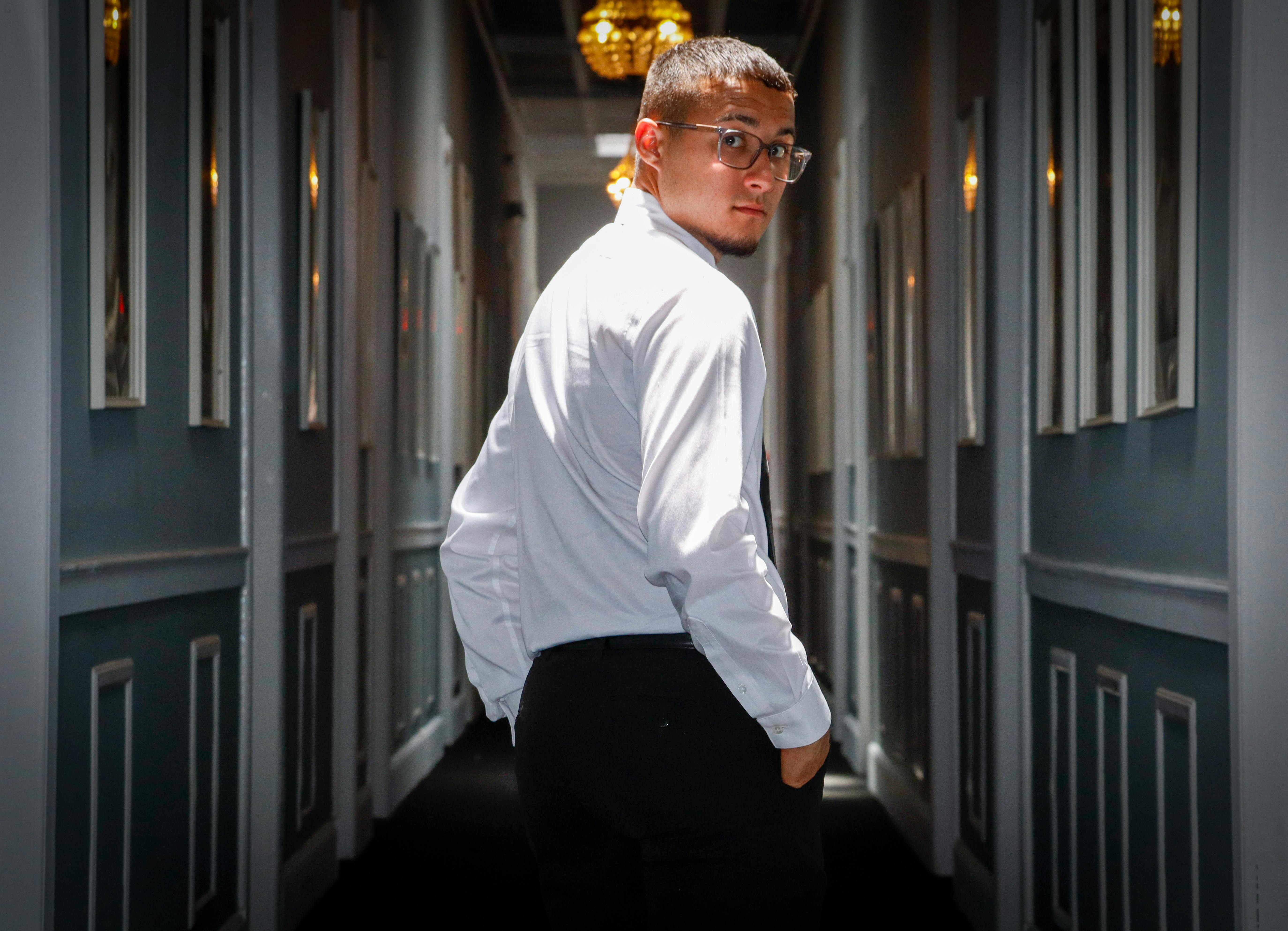 Young man in white dress shirt walking confidently through a dimly lit corridor with chandelier lighting.