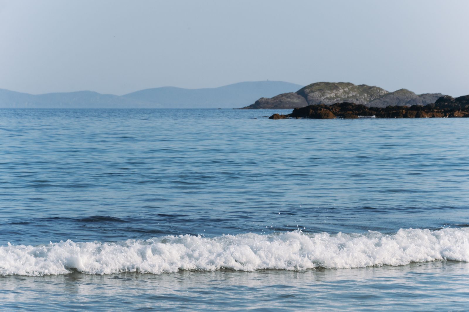 Scene of waves rolling onto the shore with distant islands under a clear sky in Ireland