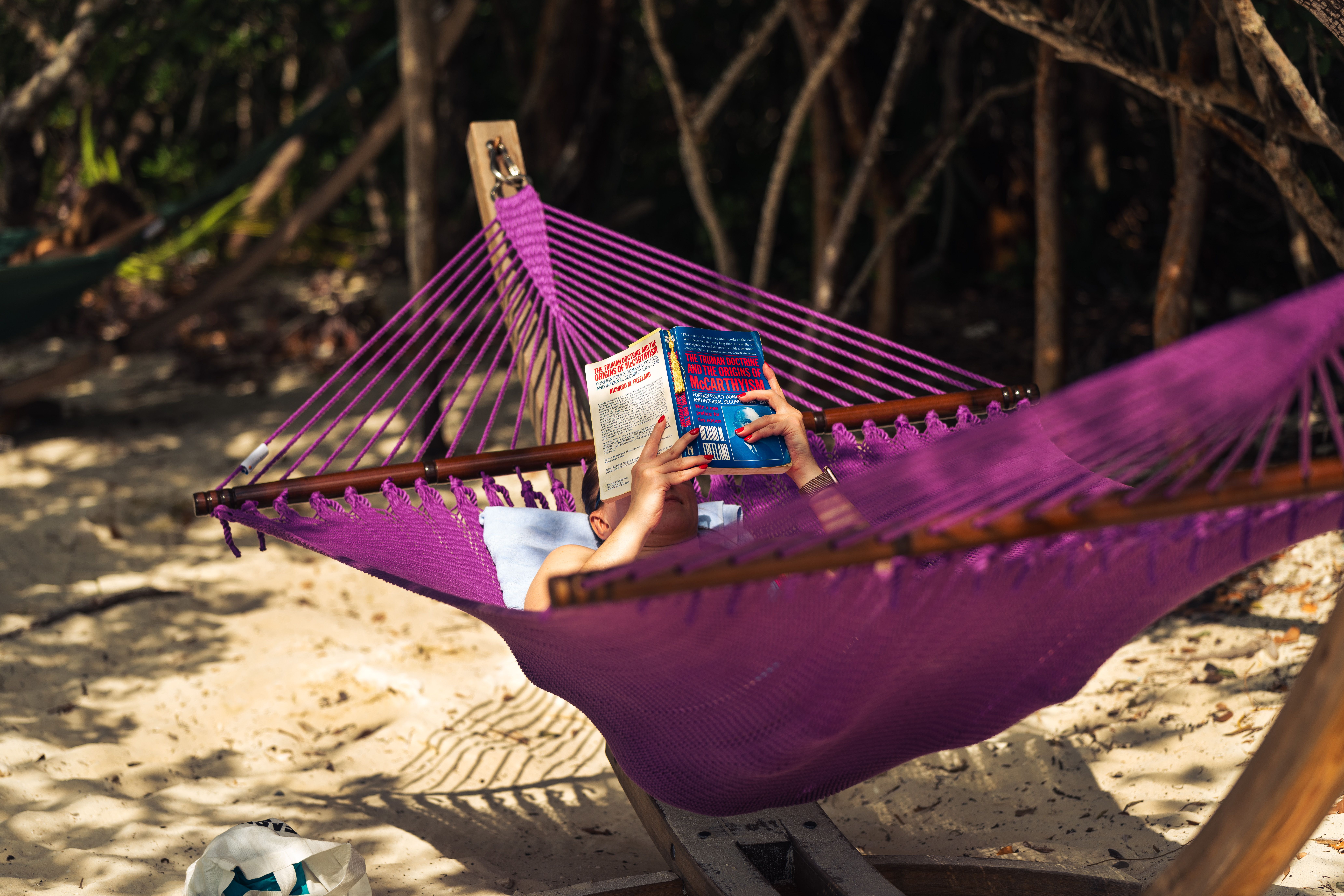 A person enjoys a peaceful read in a hammock under tropical trees on a sandy beach.