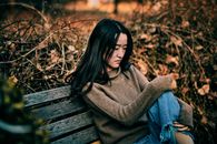 一名亚洲妇女若有所思地坐在公园的木凳上，四周散落着干树叶，神情中透出沉思