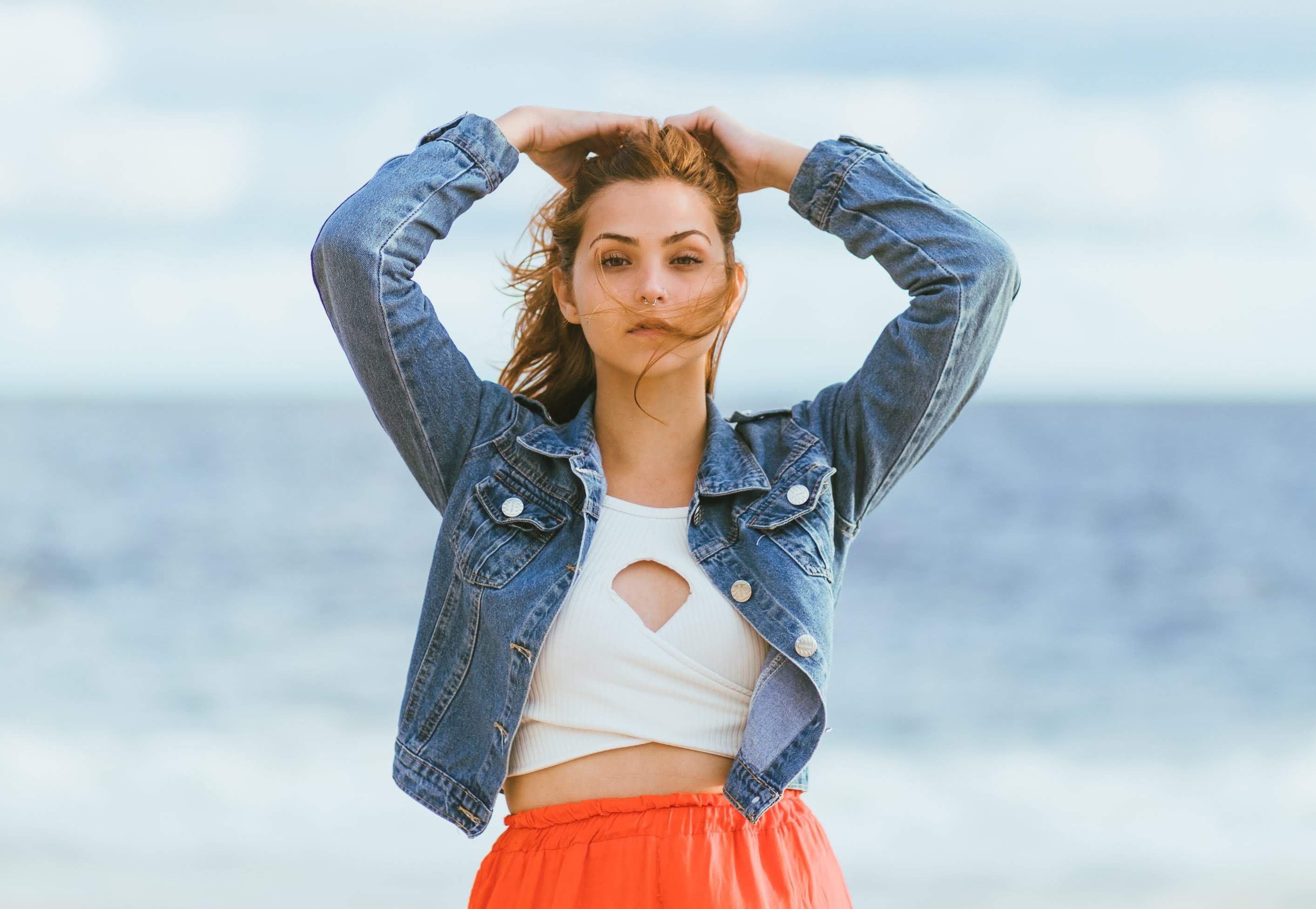 Stylish woman poses at the beach wearing a denim jacket with the ocean as a backdrop.