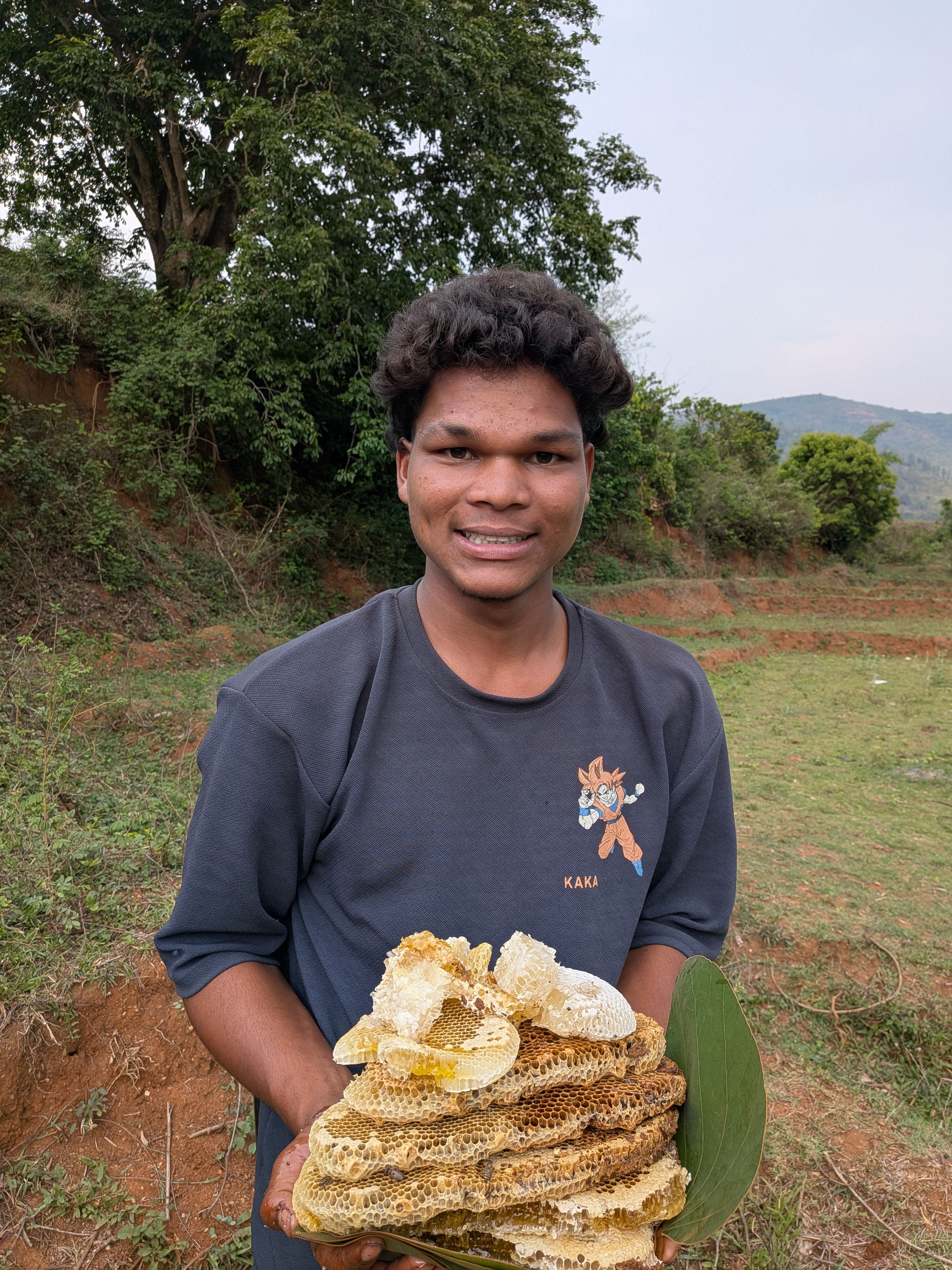 Smiling young man outdoors holding a large honeycomb on a grassy field.