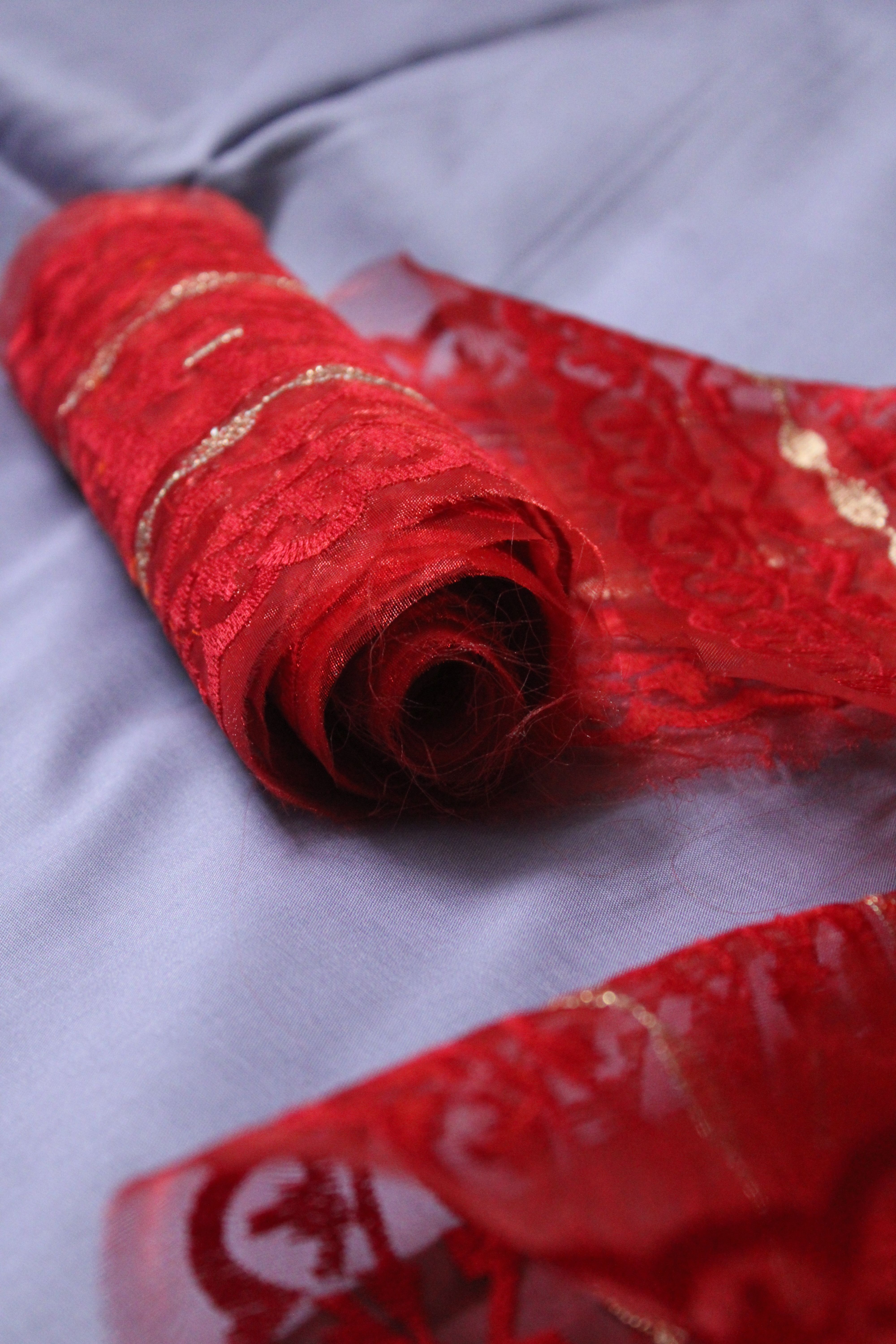 A detailed view of red lace fabric with gold accents rolled on soft blue cloth.