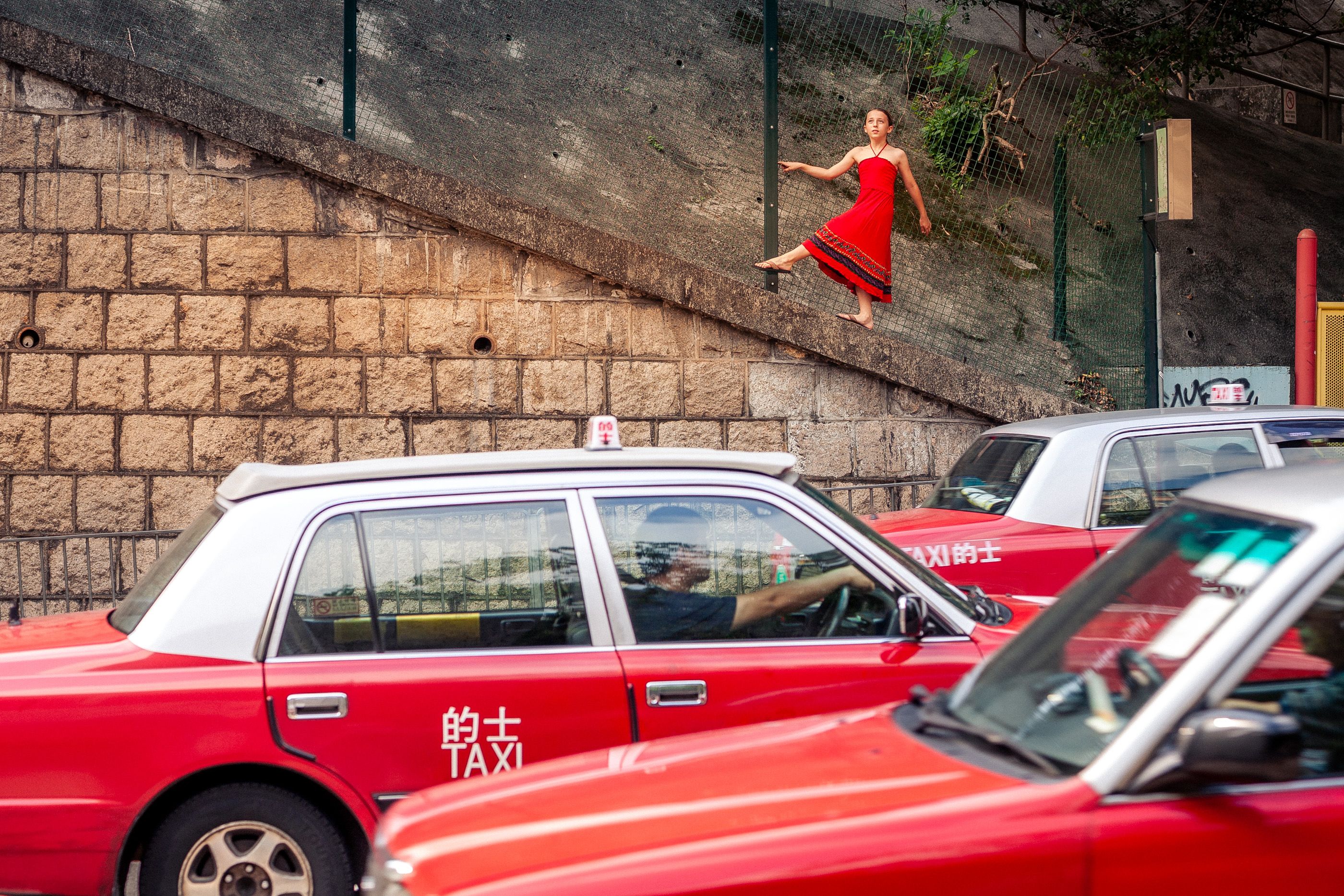 Creative street photo of a dancer in red posing above taxis in Hong Kong, showcasing urban movement.